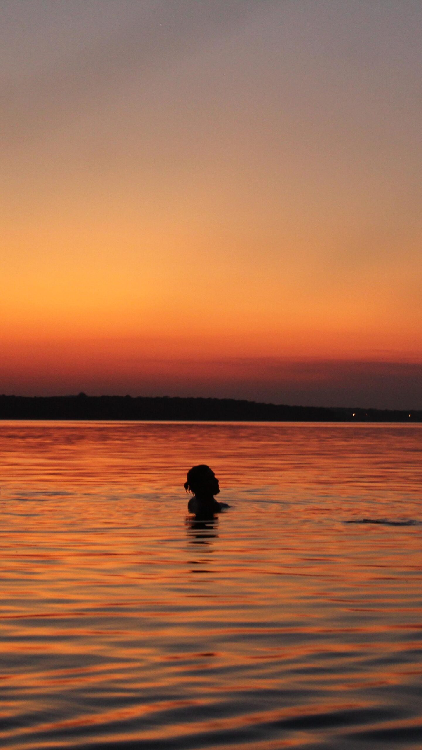 Submerged in liquid gold, the river holds everything. A person swimming in a vast river at dusk, silhouetted against a deep orange sunset sky on an Amazon cruise from Alter do Chão