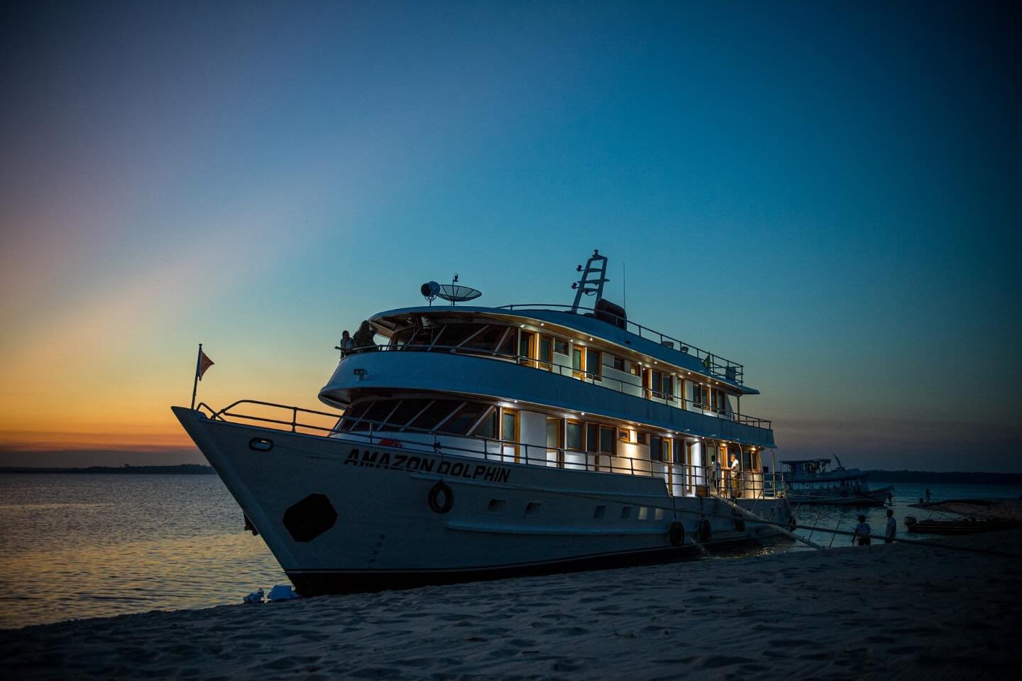 Cabin lights on, sky fading — the Amazon Dolphin settles in for the night. The Amazon Dolphin cruise vessel moored on a river sandbar at dusk, warm cabin lights glowing against a deep blue and orange sunset sky on an Amazon cruise from Alter do Chão