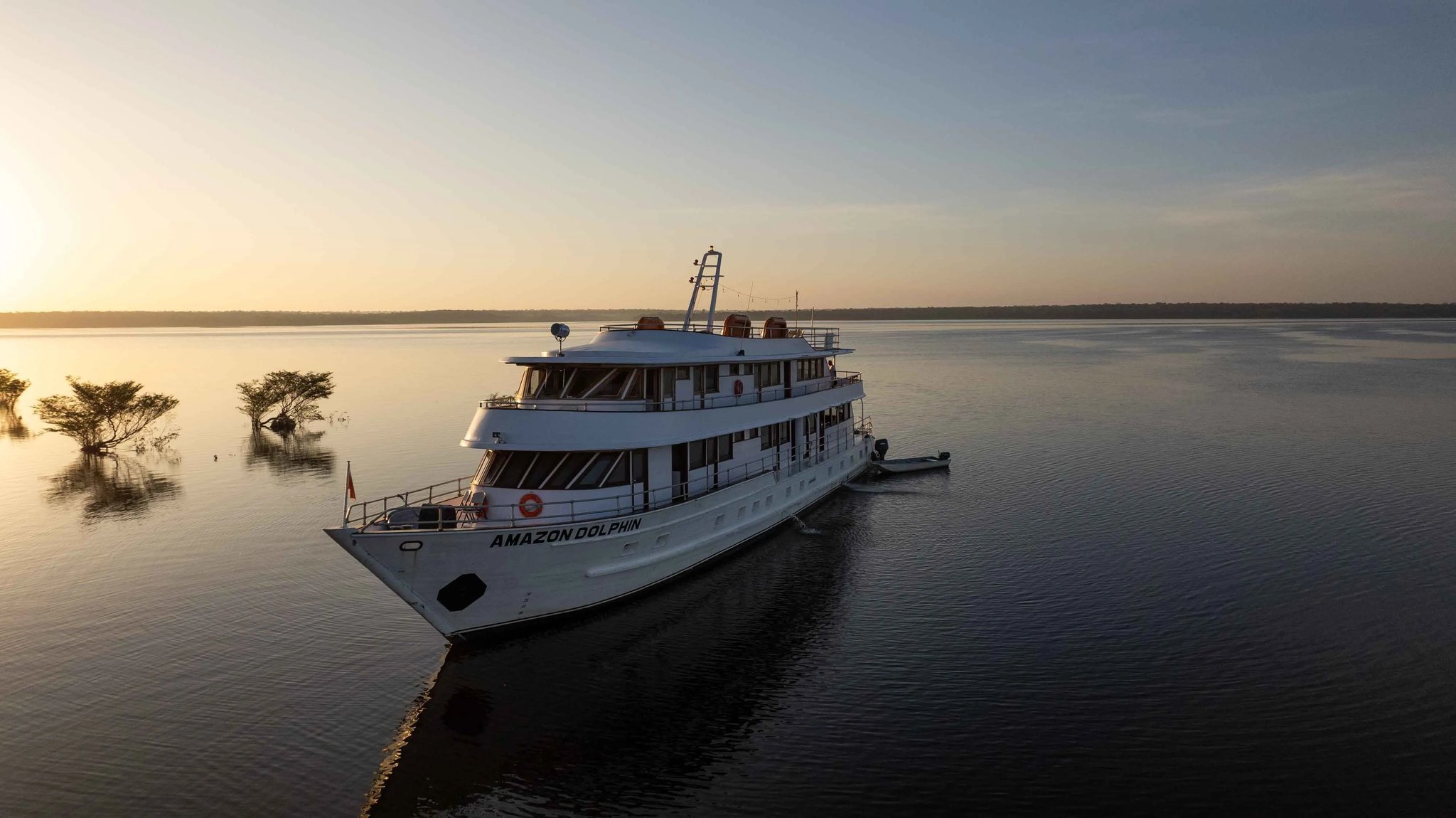 Mirrored water, a burning horizon — the Amazon Dolphin settles in for the night. The Amazon Dolphin vessel anchored on a glassy river at golden-hour sunset, part of the Amazon cruise from Alter do Chão fleet, with flooded shoreline trees silhouetted against a warm horizon