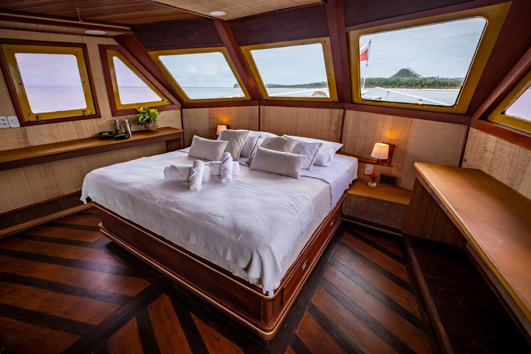 Spacious master cabin aboard an Amazon cruise from Alter do Chão, with a king-size bed dressed in crisp white linen, towel-folded animals, warm timber panelling, and panoramic windows overlooking a forested riverbank