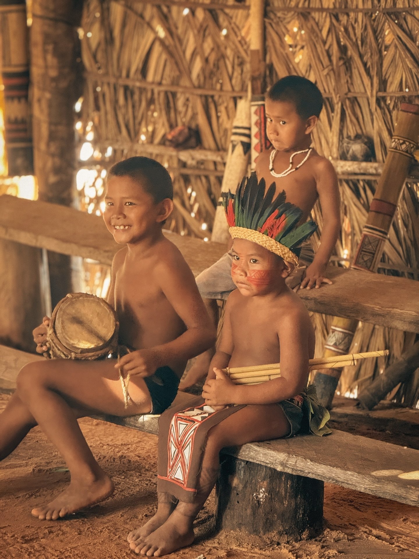 Small hands, big traditions — children keeping their culture alive. Three indigenous children in traditional face paint, feather headdress and woven accessories sit inside a thatched maloca during a cultural visit on an Amazon cruise trip in Brazil