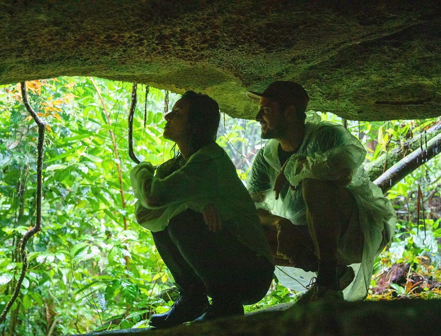 Rain gear on, heads ducked — the 700-million-year-old Madadá Cave demands respect. Two travellers crouch beneath a low sandstone overhang inside a cave on an Amazon Cruise Trip in Brazil, surrounded by lush rainforest vegetation visible through the opening