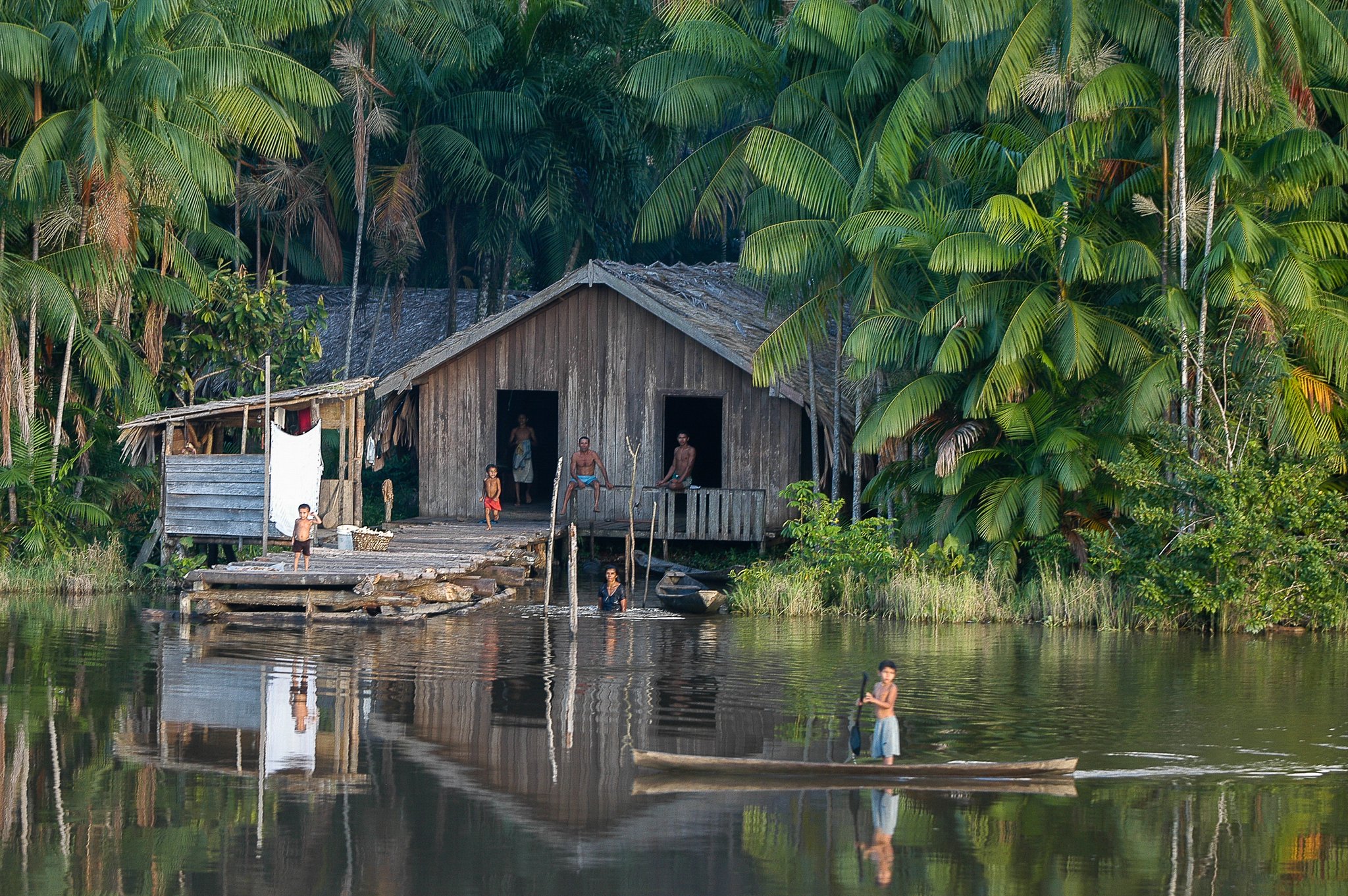 Rippled reflections and bare feet — river life unfolding at the forest's edge. Photo: J Brarymi A weathered wooden stilt house beside a still river mirrors palm-dense rainforest; a child paddles a dugout canoe in the foreground during an Amazon cruise trip in Brazil