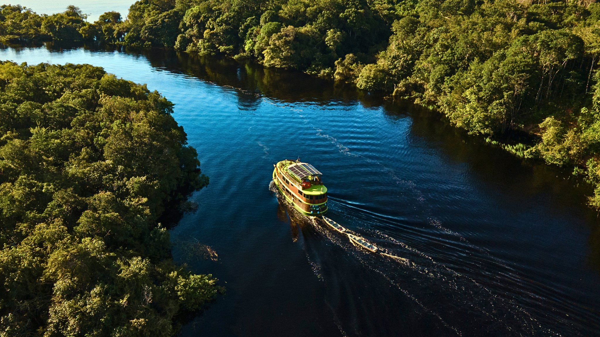 Solar panels, black water, unbroken green — deep into the Amazon. Aerial view of a solar-panelled expedition vessel on an Amazon river cruise, towing a small skiff through dark blackwater flanked by dense rainforest canopy