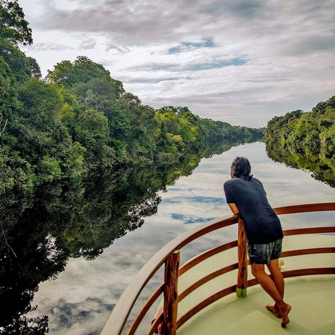 Still water, endless green — the river opens ahead. A traveller leans on the wooden bow railing of a riverboat during an Amazon jungle cruise Brazil, gazing along a mirror-still forest canal lined with dense rainforest canopy