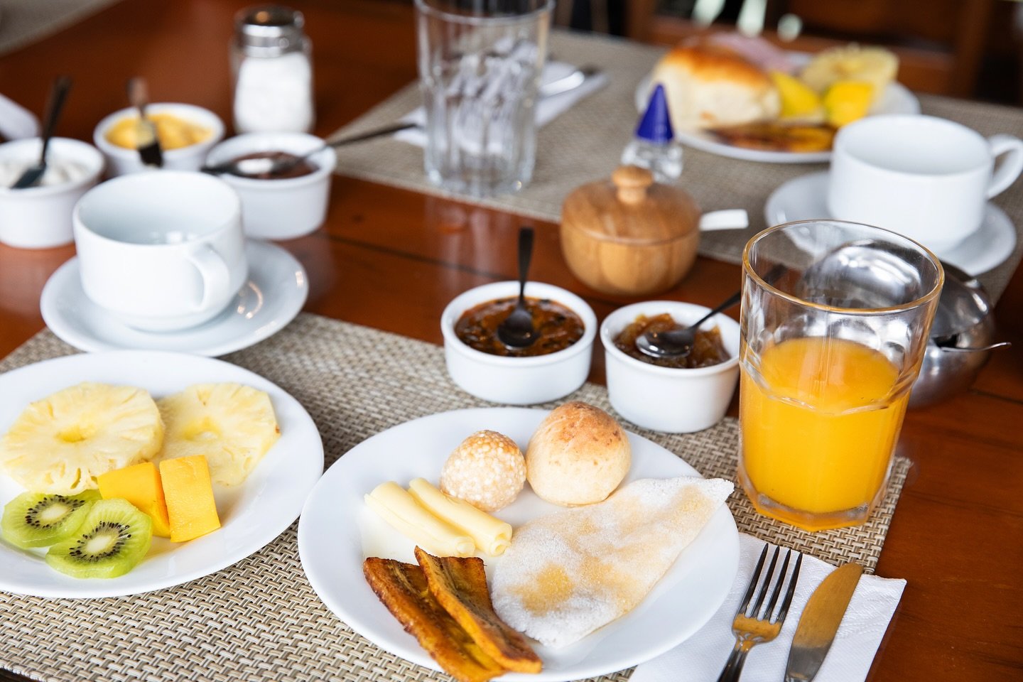 Tropical fruits, warm cheese rolls, and tapioca — fuel before the forest. A full Amazon jungle cruise Brazil breakfast spread with pão de queijo, tapioca, fried plantains, sliced cheese, fresh pineapple, kiwi and mango, fruit preserves, orange juice and coffee cups set on a woven placemat