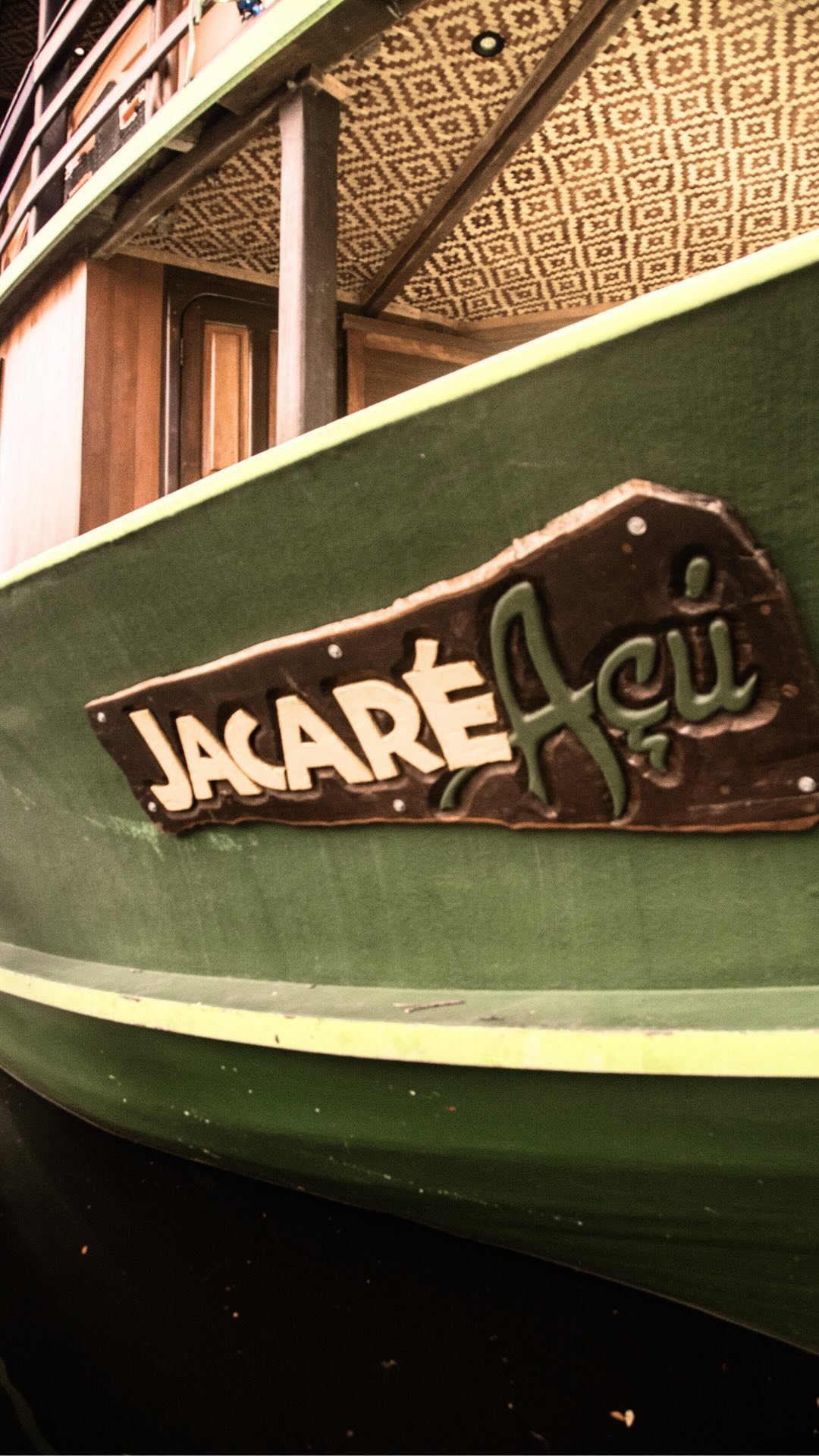 Close-up of the carved wooden nameplate reading 'Jacaré Açu' bolted to the green hull of the Amazon jungle cruise Brazil vessel, with a patterned ceiling and wooden structure visible above
