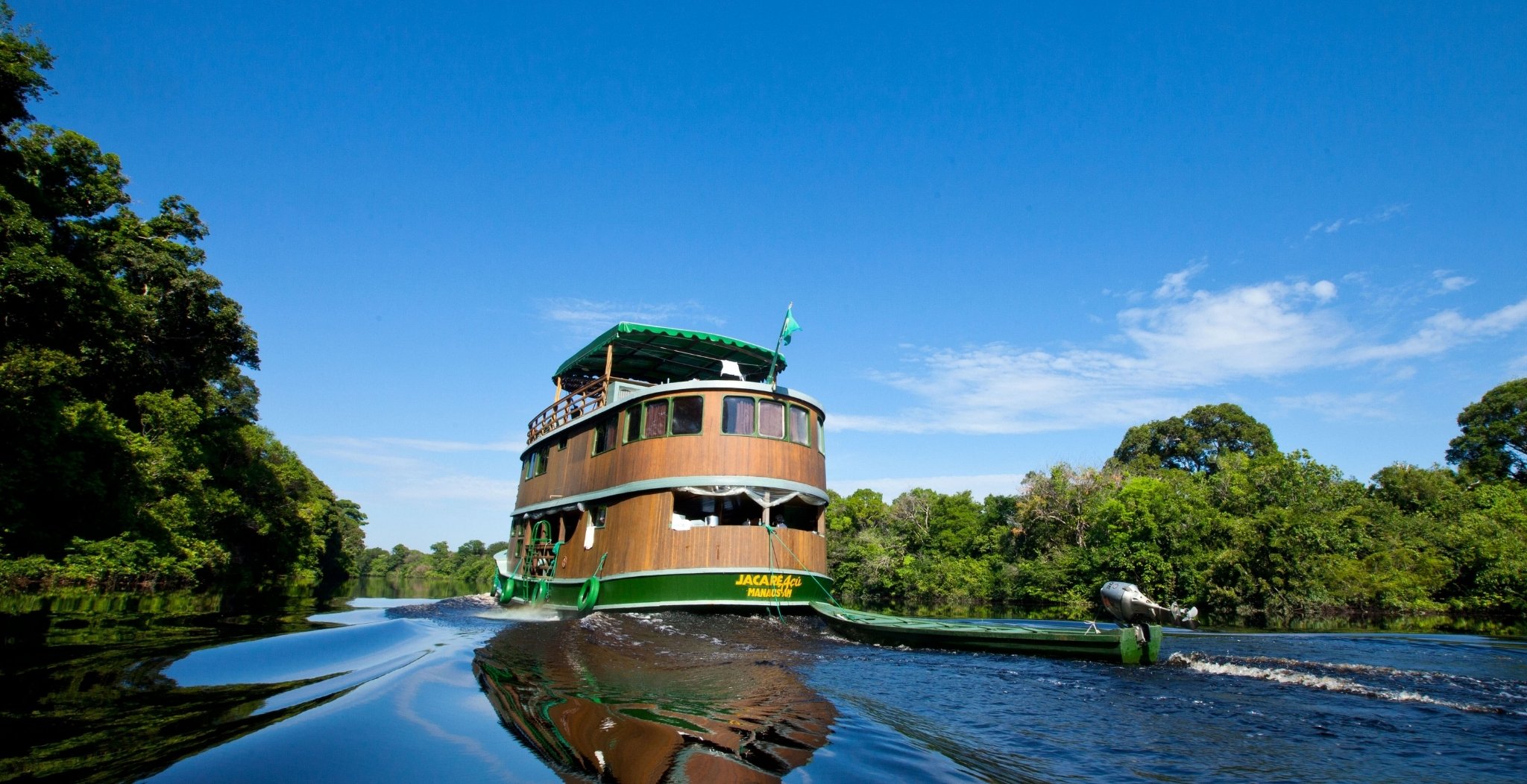 The wooden-hulled Jacaré Açu vessel cruises a calm black-water river channel on an Amazon jungle cruise in Brazil, flanked by dense rainforest canopy under a bright blue sky