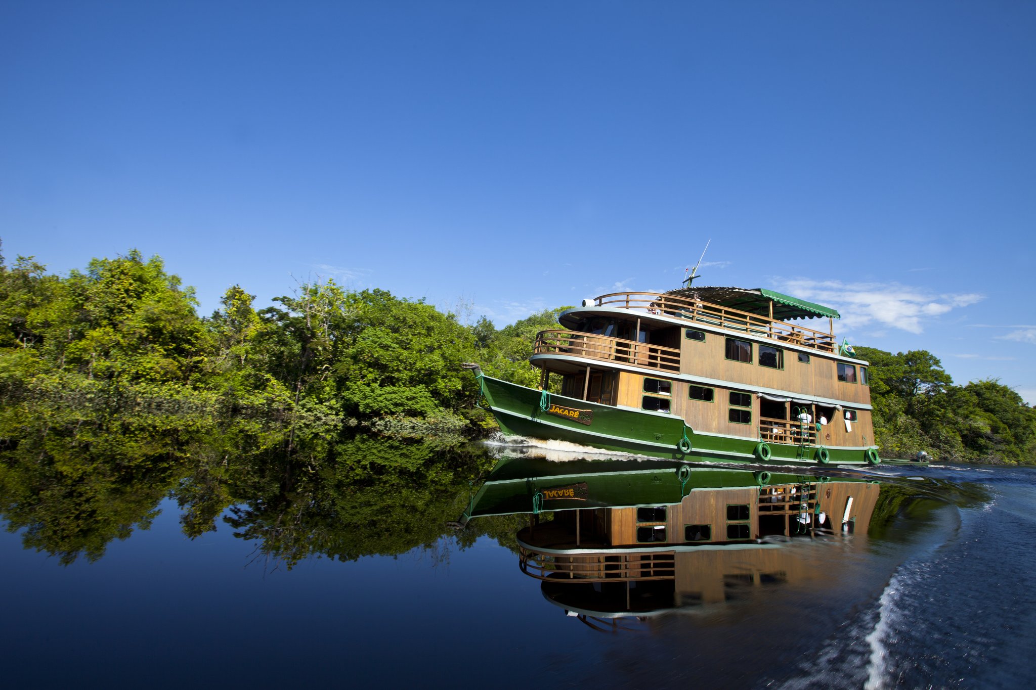 Hull and reflection, inseparable — the Jacaré glides through a still black-water channel. The wooden-hulled Jacaré riverboat cruising a dark-water Amazon jungle cruise Brazil channel, its green and timber hull perfectly mirrored in the glassy river surface, dense rainforest lining both banks under a vivid blue sky
