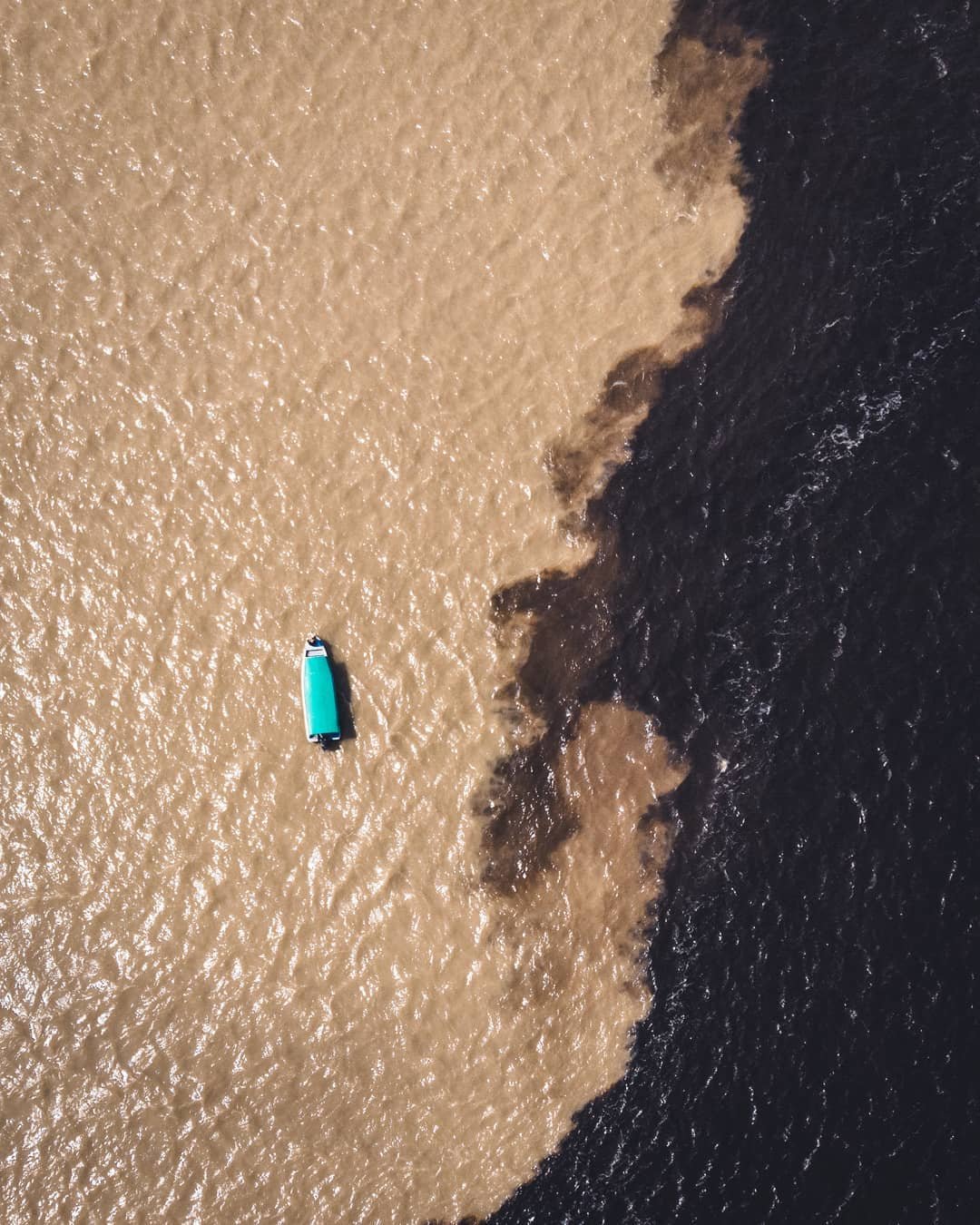 Two rivers, one frame — impossible not to stare. Aerial view of a small teal-roofed boat on an Amazon jungle cruise in Brazil, crossing the boundary where sandy brown water meets dark black water at the Meeting of the Waters