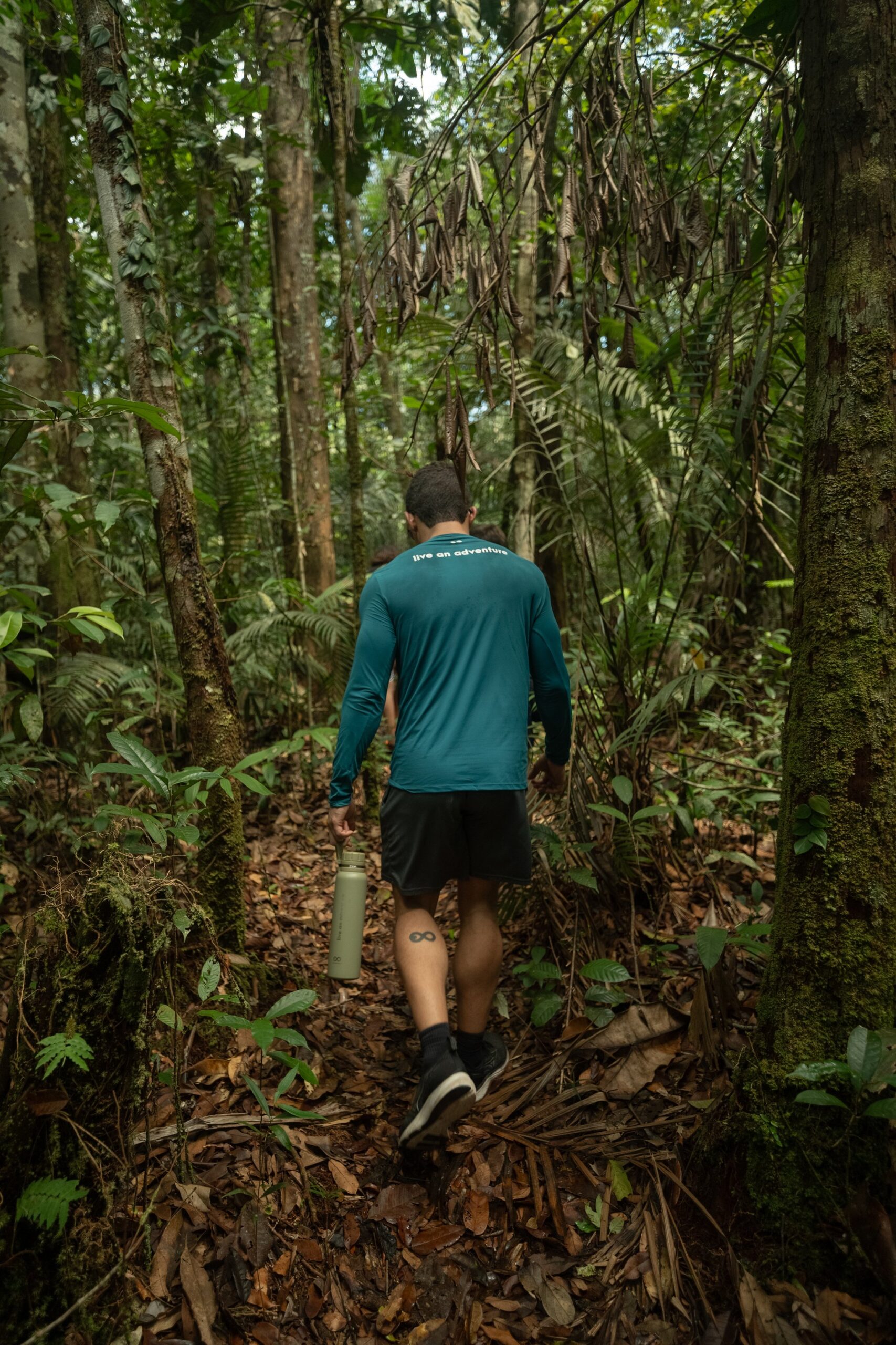 Leaf litter underfoot, canopy closing in — the forest swallows you whole. A hiker in a teal long-sleeve shirt carries a water bottle along a narrow trail through dense primary rainforest during an Amazon jungle cruise Brazil excursion