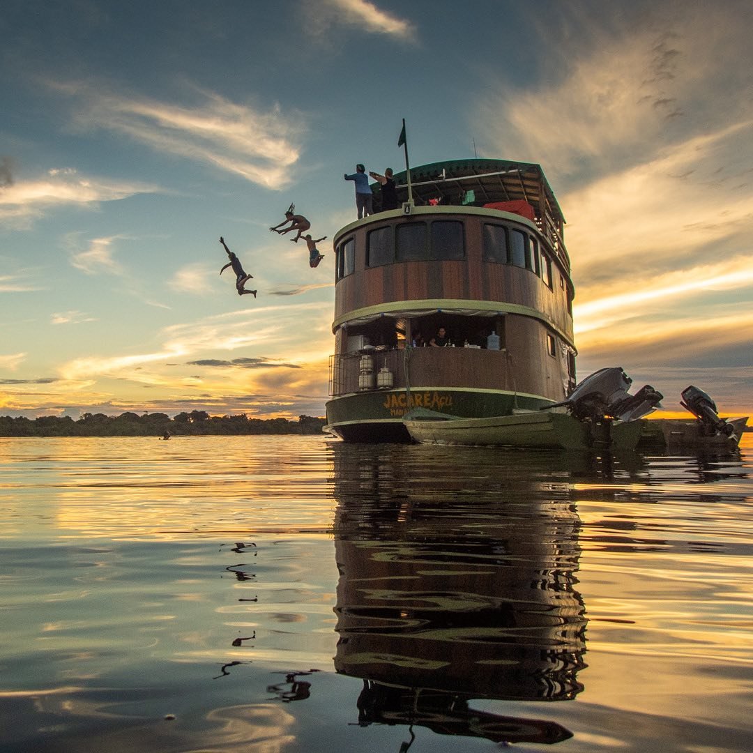 Leap, splash, repeat — sunset dips off the top deck. Passengers leaping from the upper deck of the Jacaré Açu riverboat into a glassy river at golden-hour sunset on an Amazon jungle cruise Brazil