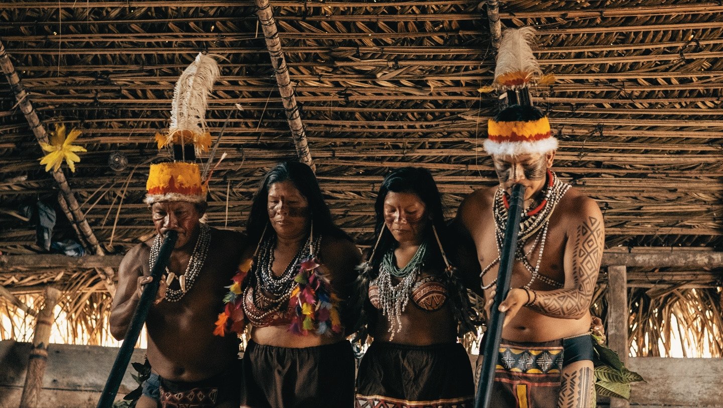 Vivid pigments, layered beads, and feathers earned through tradition — a Tuiuka ceremony like no other. Four Tuiuka community members in traditional feathered headdresses, body paint, and beaded necklaces performing a ceremony inside a thatched-roof maloca during an Amazon jungle cruise Brazil cultural visit