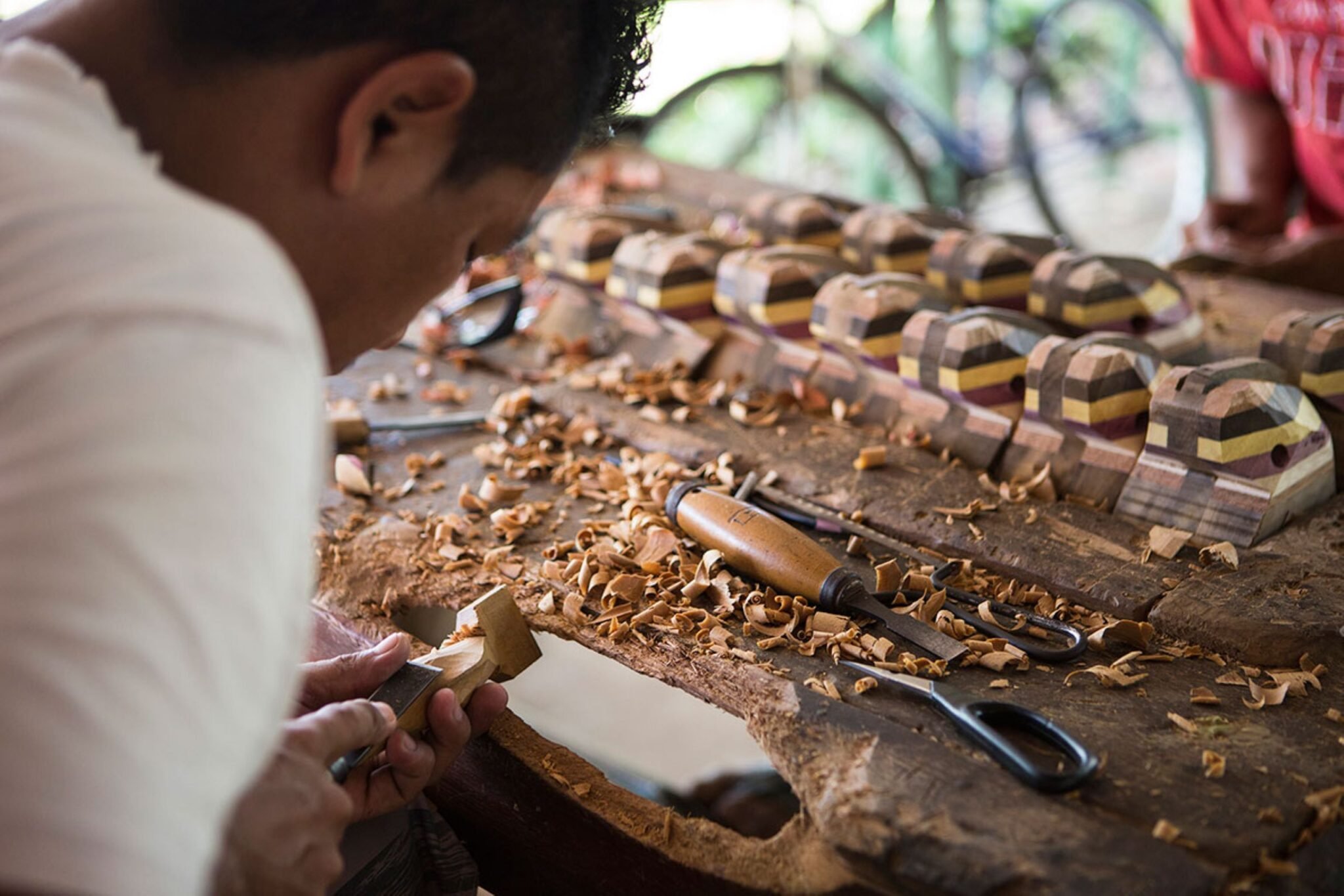 Shavings, chisels, and hours of patience — local craftsmanship up close. A local artisan carves a small wooden piece by hand on a workbench scattered with curled wood shavings, with a row of finished multi-toned laminated wood crafts displayed behind him — a cultural highlight of an Amazon jungle cruise Brazil itinerary