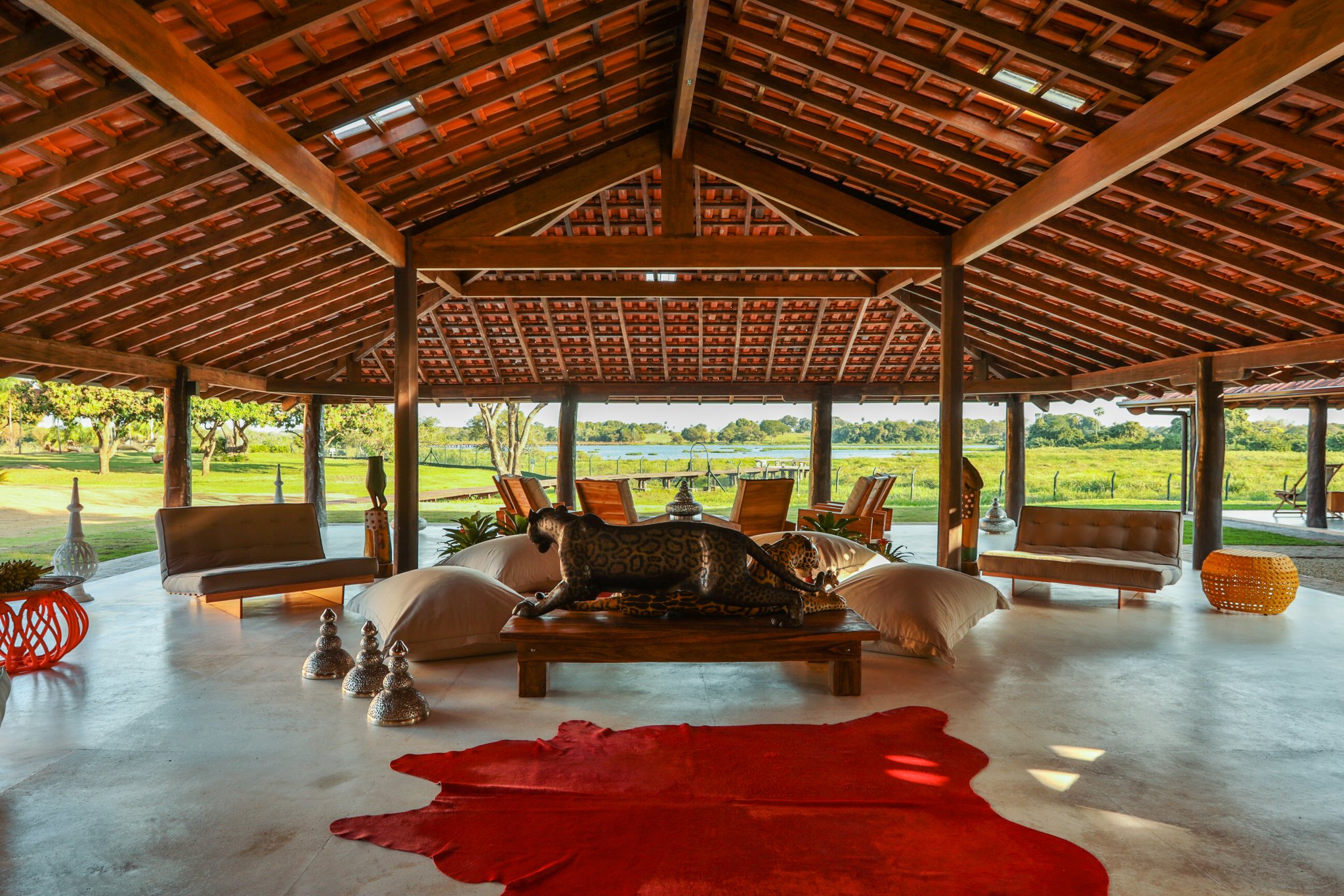 Open-air lounge of an amazon jungle lodge in Brazil featuring a soaring timber roof, jaguar sculpture centrepiece, floor cushions, and sweeping wetland views beyond the open pavilion