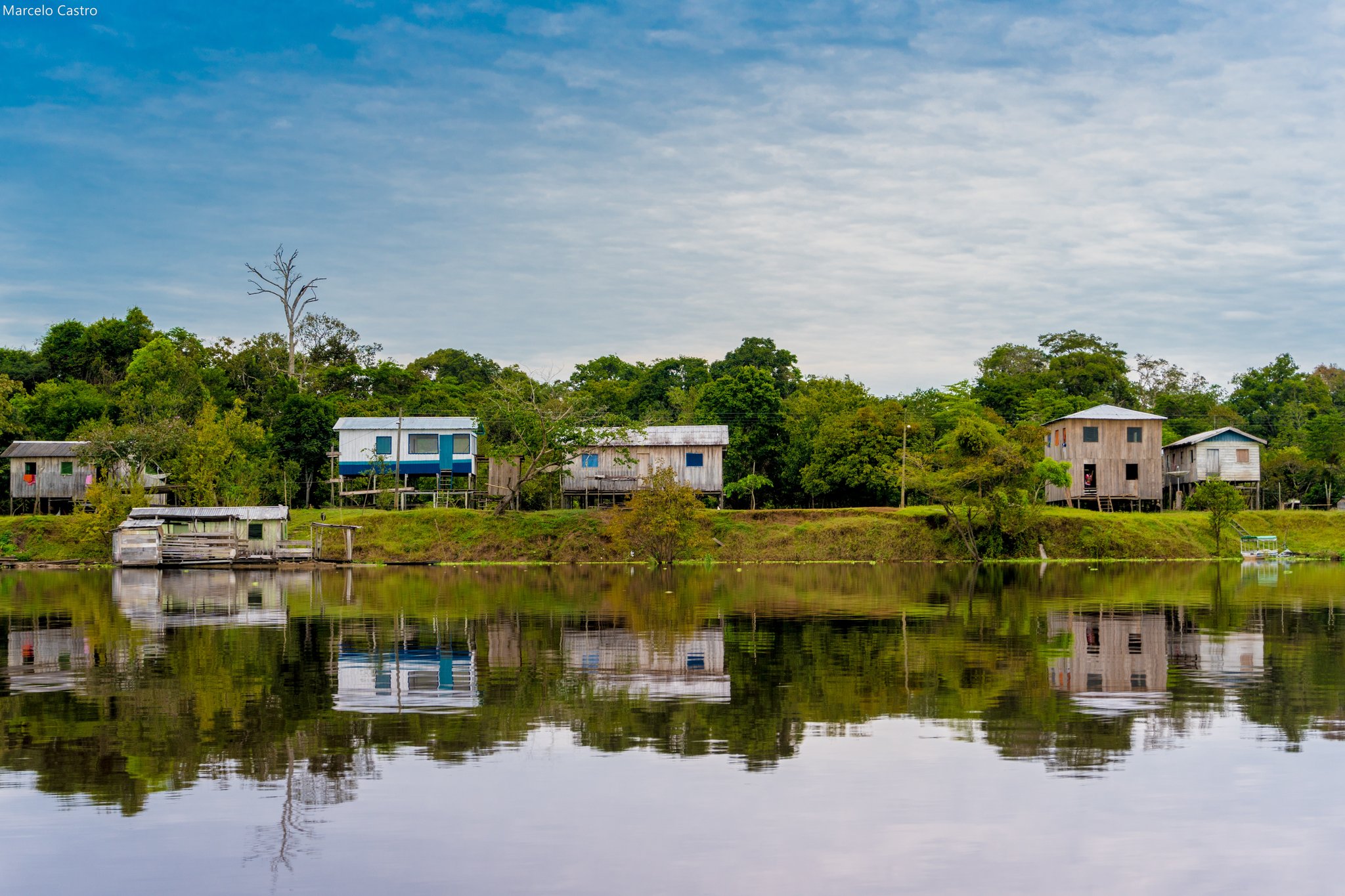 Wooden stilt houses reflected in a calm river channel, with dense rainforest canopy behind them — a riverside community seen during an amazon rainforest ecolodge trip in brazil