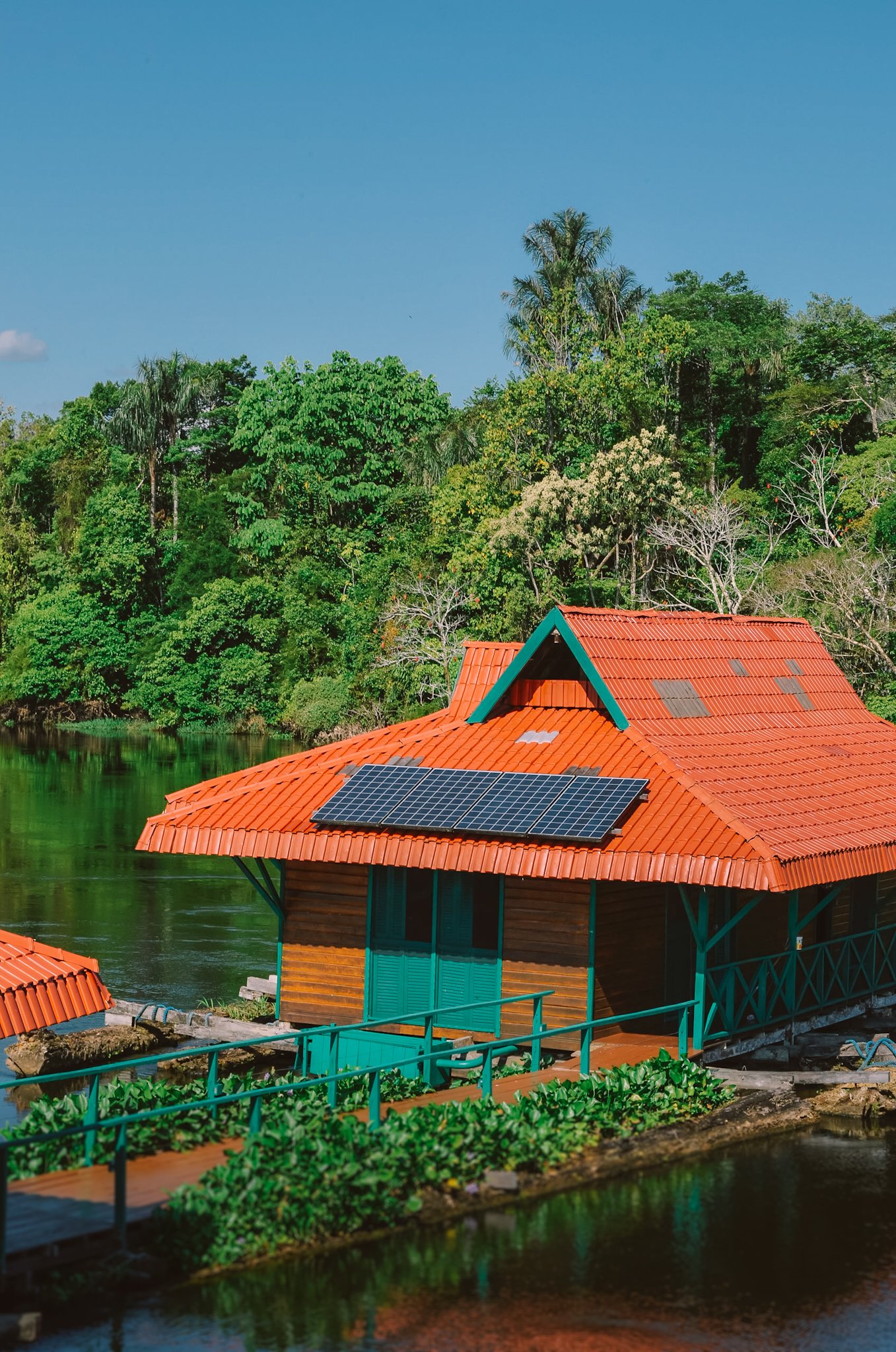 A solar-panelled wooden cabin with a vivid orange roof floats on a dark river during an amazon rainforest ecolodge trip in brazil, surrounded by dense tropical canopy under a clear blue sky