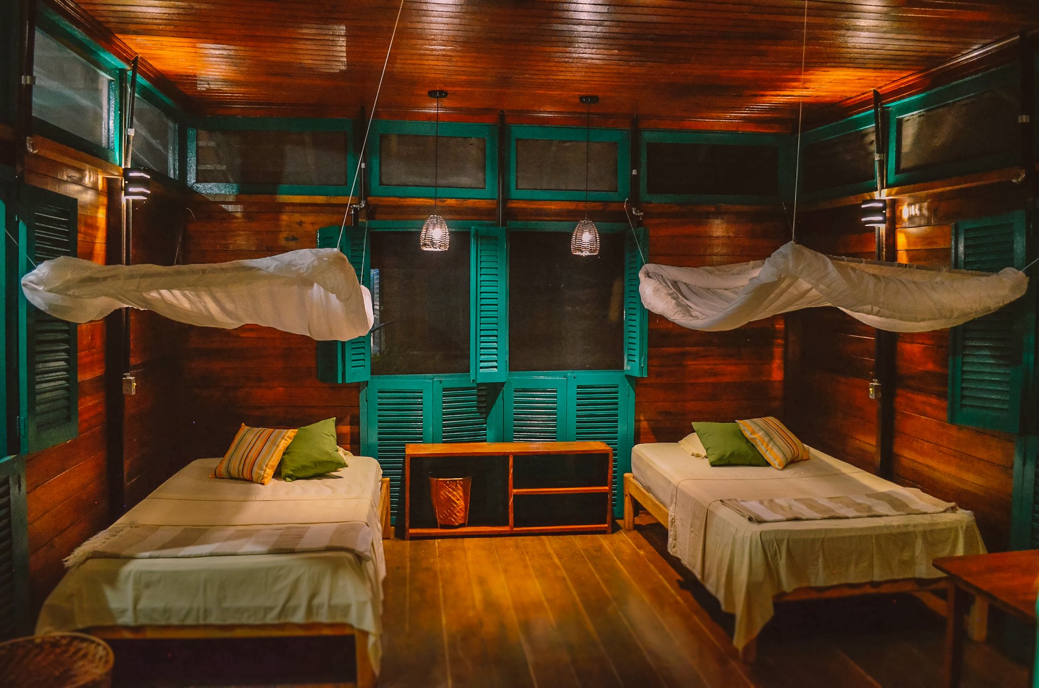 Twin-bed room at an amazon ecolodge brazil with suspended mosquito nets, warm hardwood walls, teal shuttered windows, and woven pendant lights