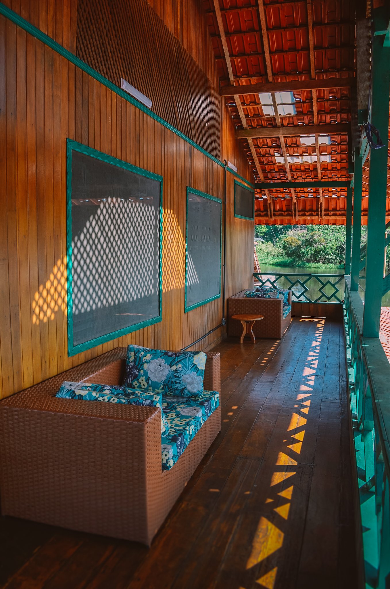 A shaded wooden veranda of an amazon rainforest ecolodge trip in brazil property, with wicker seating and tropical-print cushions, dappled sunlight casting lattice shadows across the hardwood floor, and a green riverine landscape visible at the end of the corridor