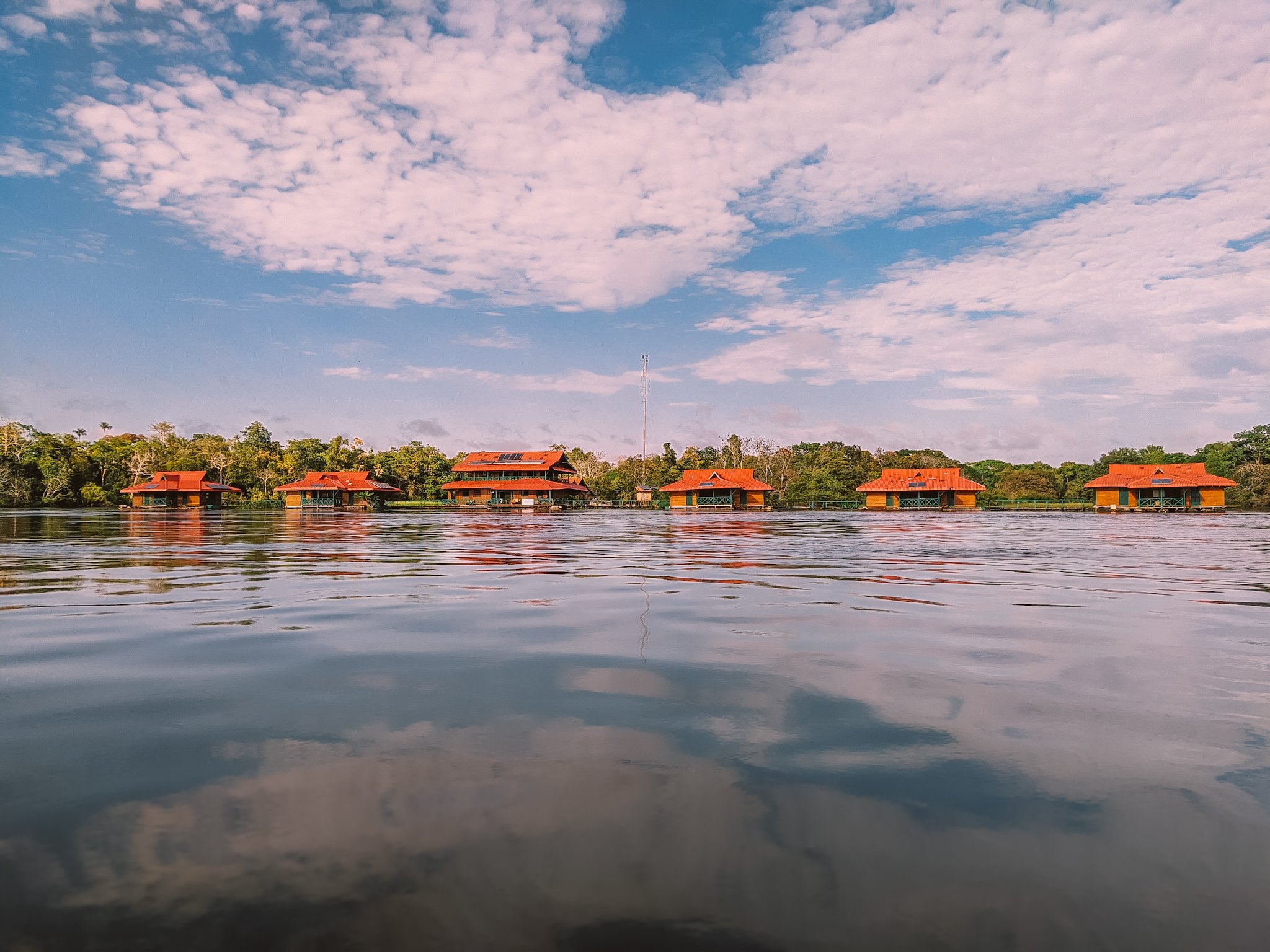 Red-roofed wooden bungalows of an amazon rainforest ecolodge trip in brazil seen from water level, with clouds reflected in the calm river and dense tropical forest behind