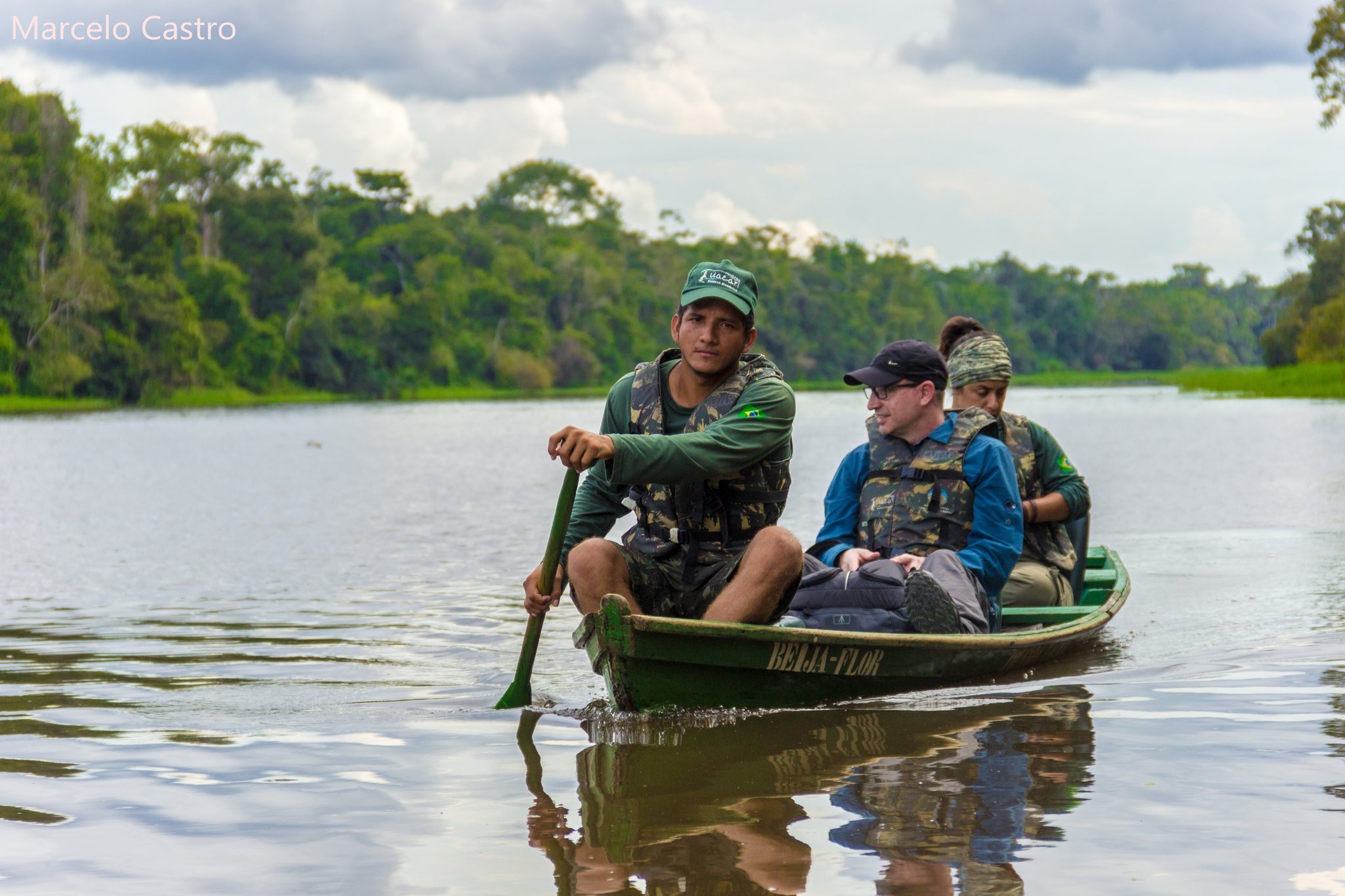 A local guide paddles a wooden canoe named Beija-Flor carrying two ecotourists across a calm river channel on an amazon rainforest ecolodge trip in brazil, with dense floodplain forest lining the far bank