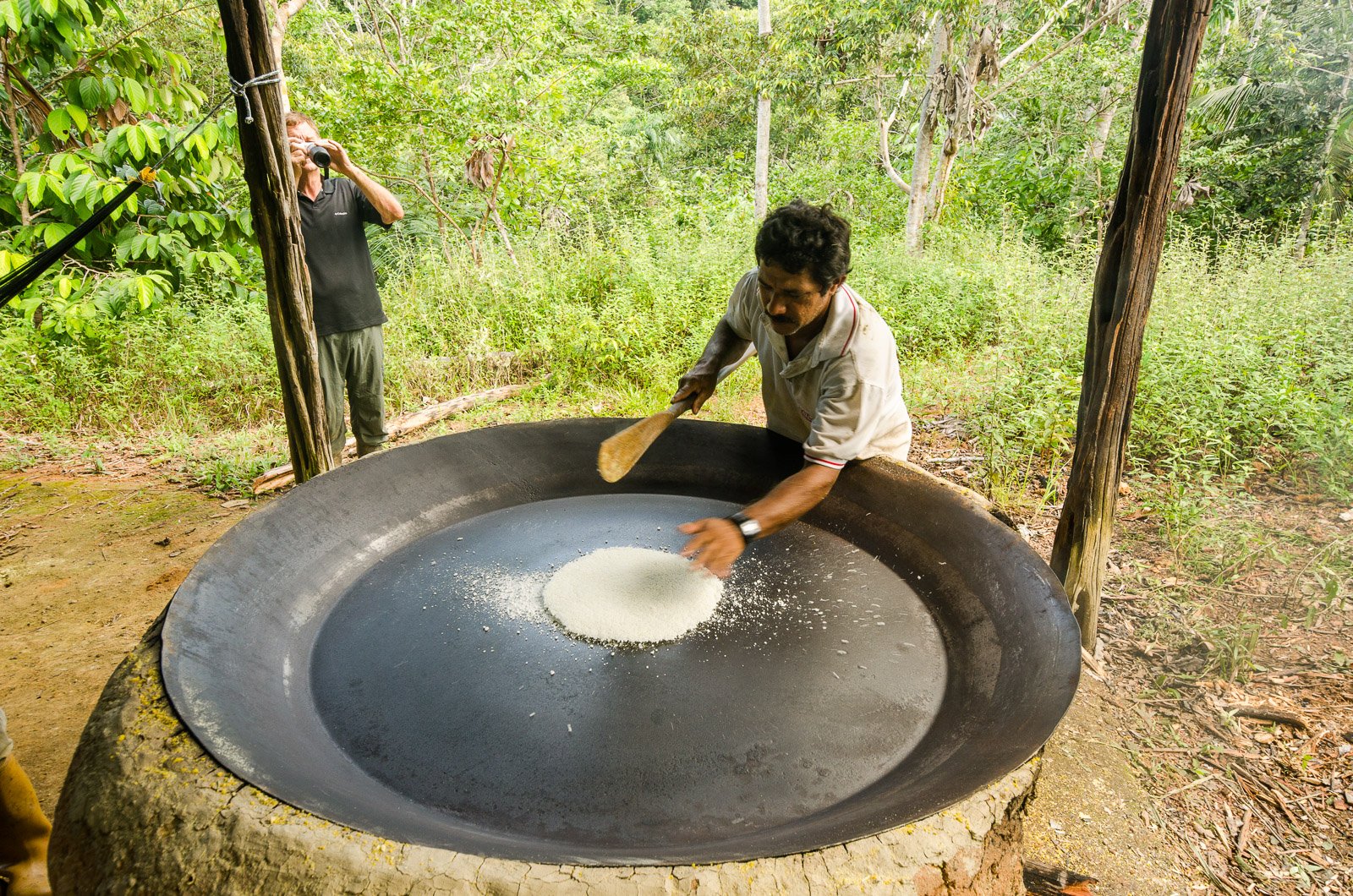 A local man toasts cassava flour on a large iron griddle over an open fire during an amazon rainforest ecolodge trip in brazil, with a visitor photographing birds in the jungle background