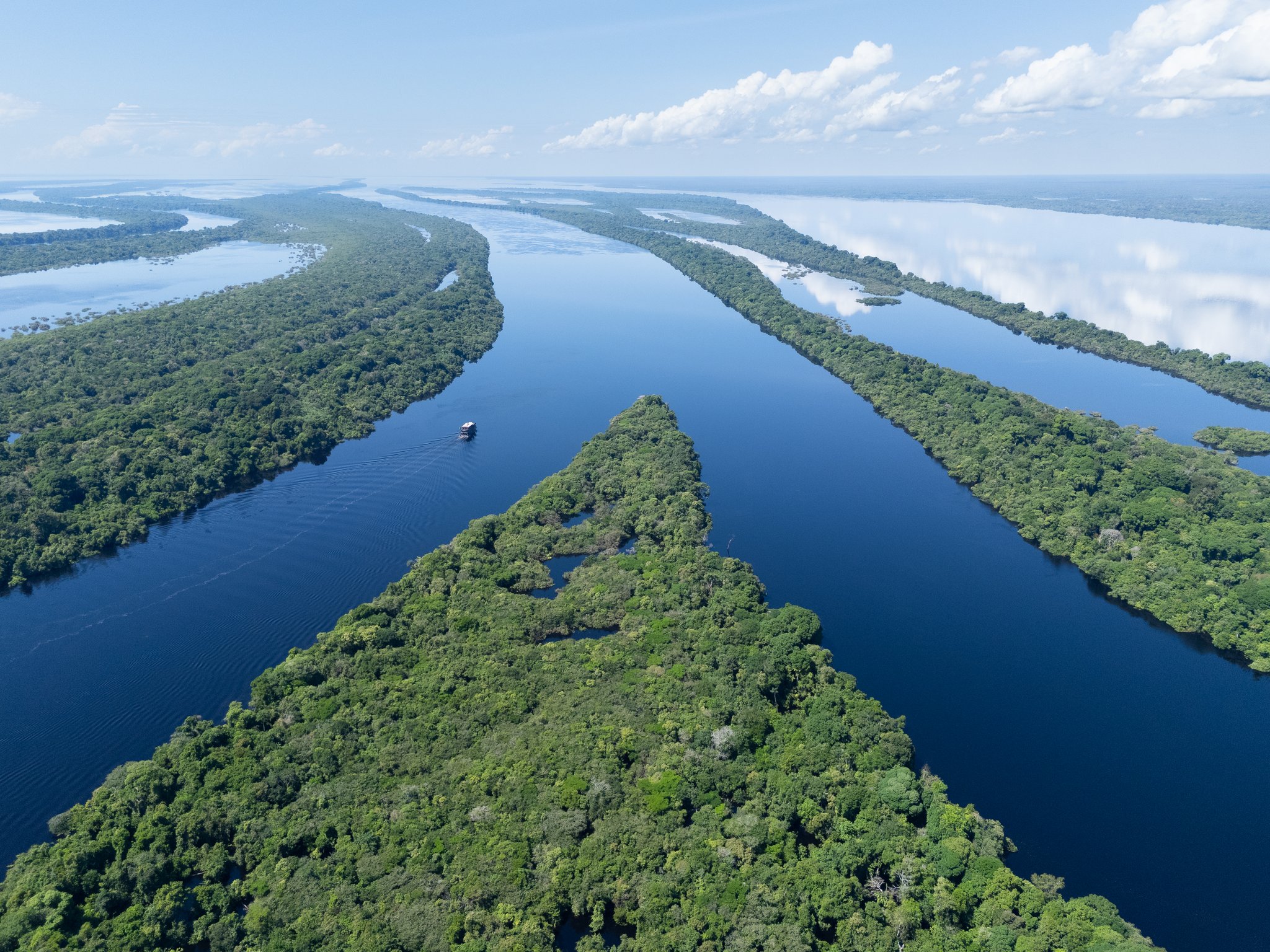 Aerial view of an Amazon River Cruise Brazil vessel navigating deep-blue blackwater channels winding between lush jungle islands of the Anavilhanas archipelago under a clear sky