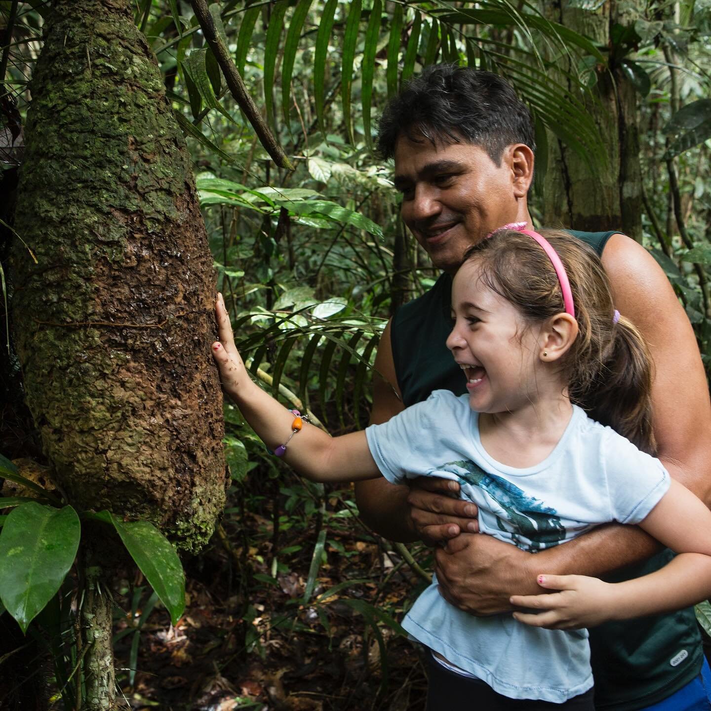 Pure joy, rough bark, and a forest that's older than memory. A smiling girl touches the textured bark of a massive rainforest tree during an Amazon River Cruise Brazil excursion, held by a laughing adult guide in dense green jungle