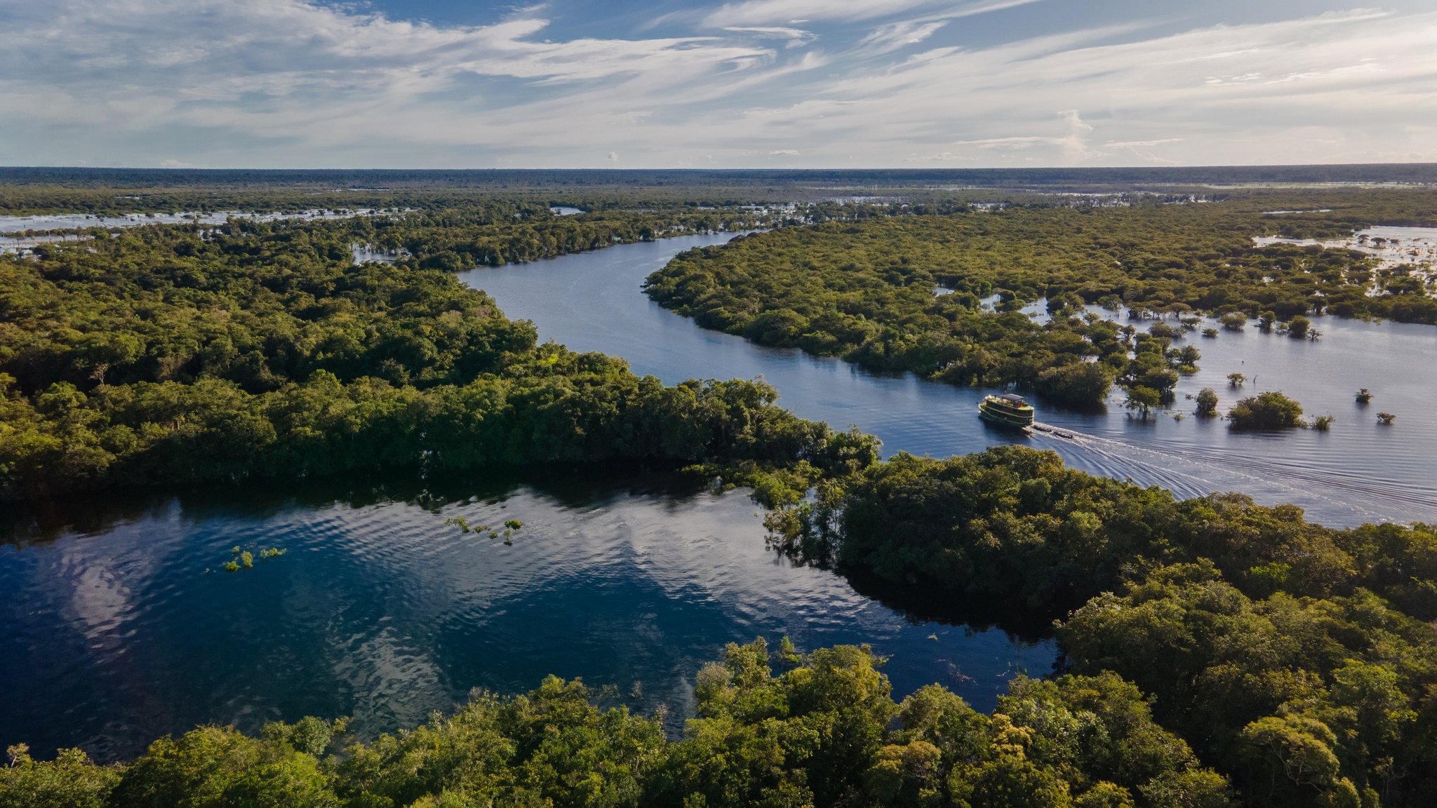 Aerial view of a small expedition vessel navigating a winding river channel through flooded Amazon rainforest on an Amazon River Cruise Brazil