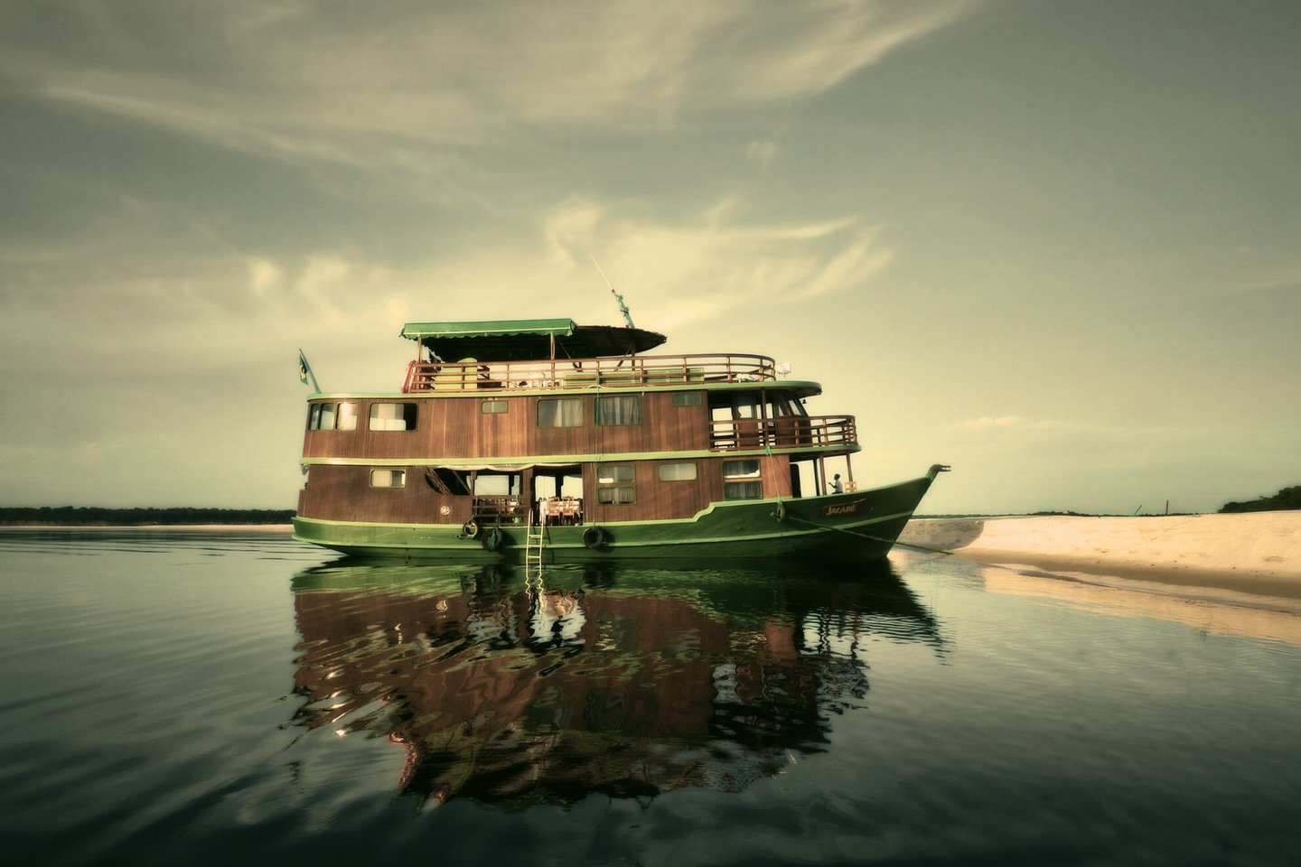 A three-deck wooden river houseboat with green trim, named Jacabé, moored beside a white sandbar on a calm dark river during an Amazon River Cruise Brazil expedition