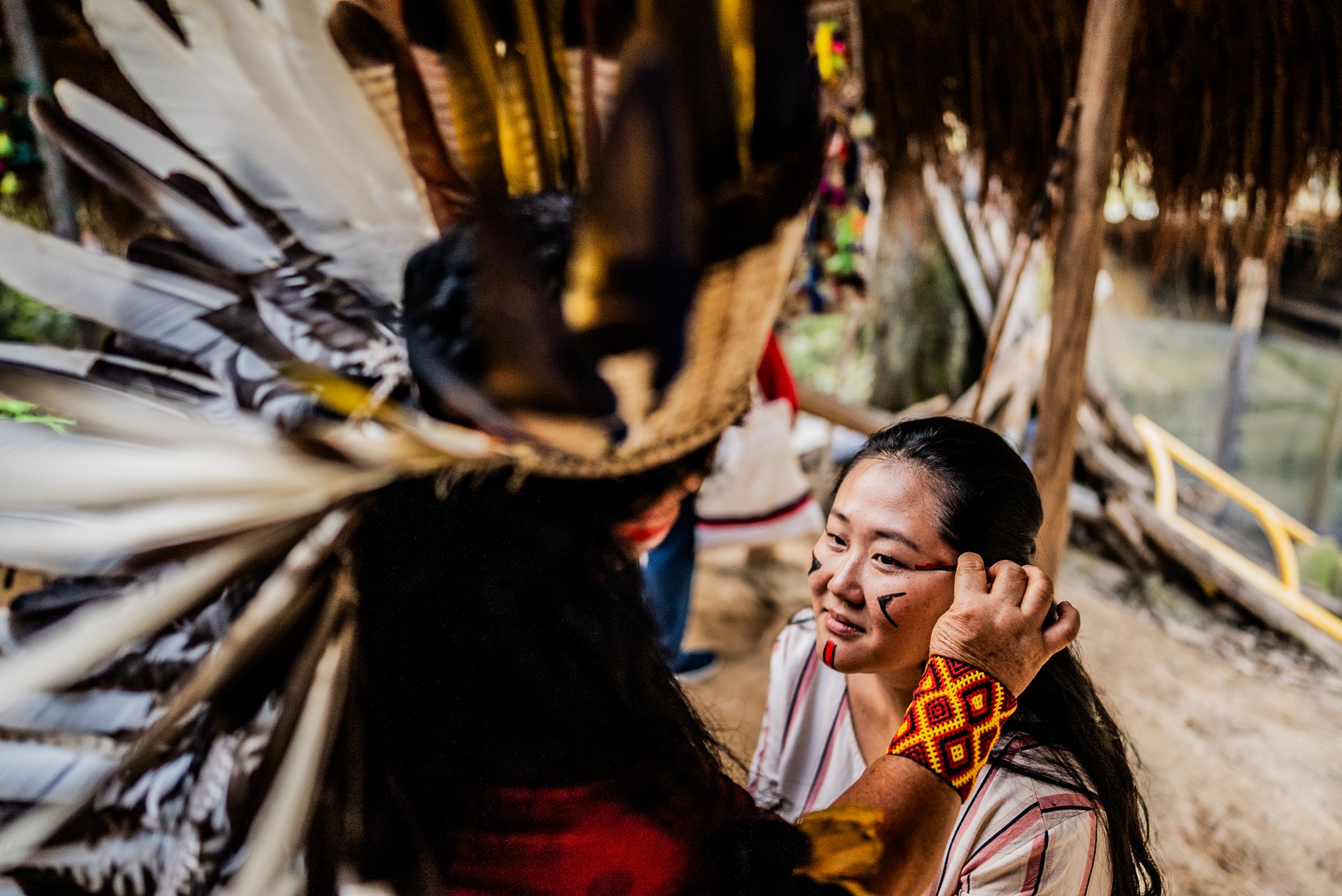 Geometric lines, slow hands, and a story painted onto skin. Photo: FG Trade A smiling visitor receives traditional geometric face paint from an Indigenous community member wearing a large feathered headdress, during an Amazon River Cruise Brazil cultural encounter under a thatched shelter