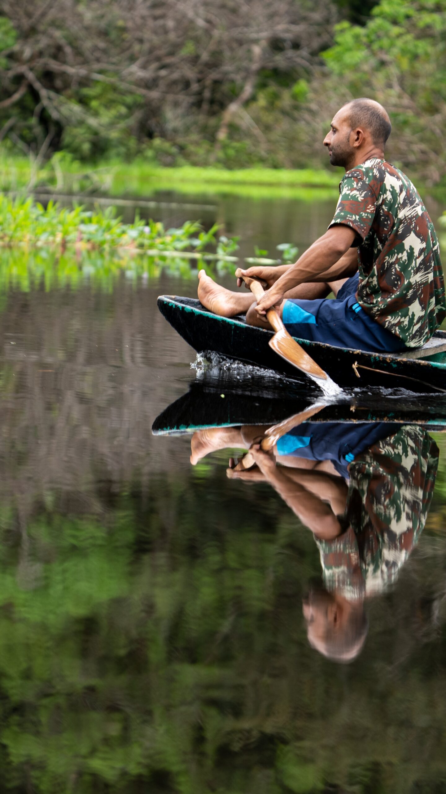 A man paddles a traditional wooden canoe across glassy, still water on an Amazon survival tour, his reflection perfectly mirrored on the dark surface beside lush green riverbank vegetation
