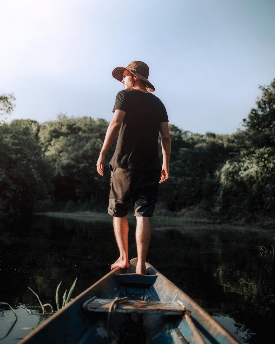 A man in a straw hat stands barefoot on the bow of a weathered wooden canoe, gazing across a calm dark river flanked by dense green forest on an Amazon survival tour