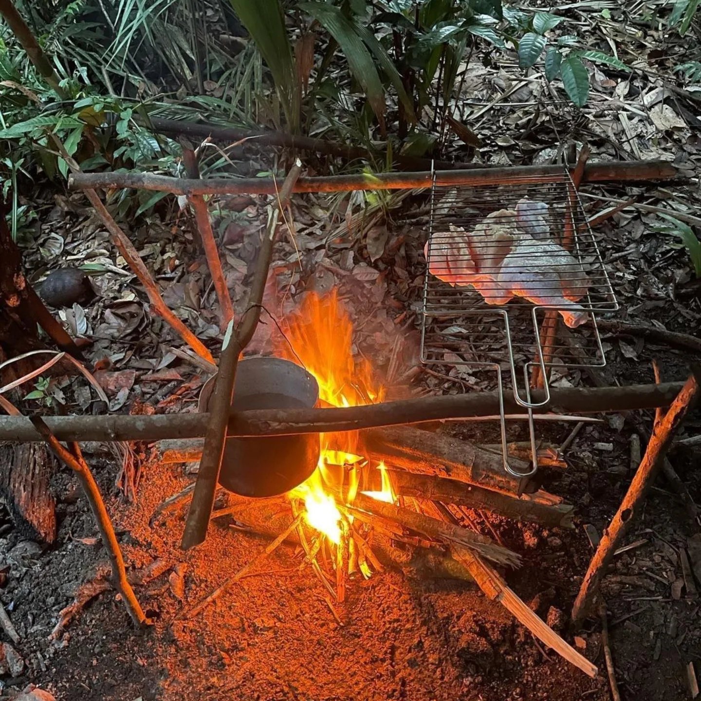 A rustic bamboo cooking frame over an open wood fire in the Amazon survival tour, with a clay pot suspended above the flames and food grilling on a wire rack, surrounded by dense jungle undergrowth