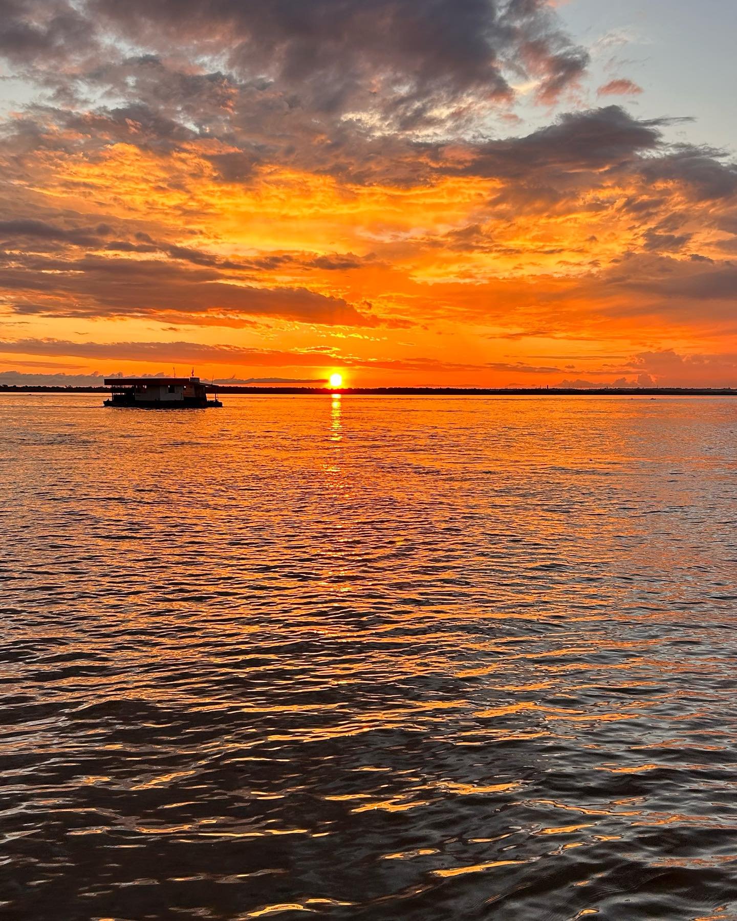 A houseboat silhouetted against a blazing orange and red sunset reflected across a wide Amazonian river on an Amazon survival tour