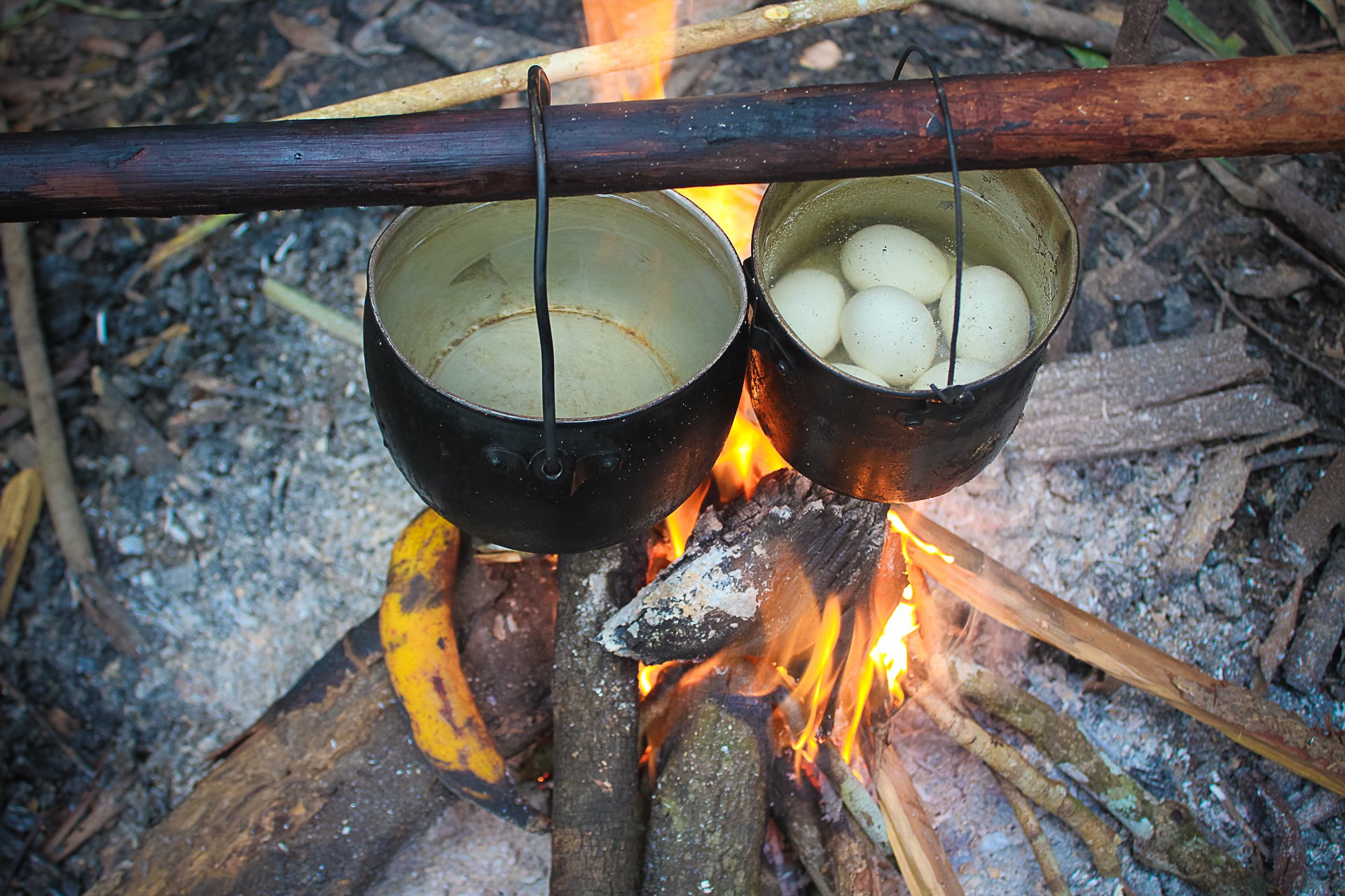 Two pots, one fire, deep jungle — the oldest kitchen in the world. Photo: Joshua Lacruz Two blackened iron pots hanging over an open wood fire on a rustic stick frame during an Amazon trek and survival tour, surrounded by dense tropical vegetation