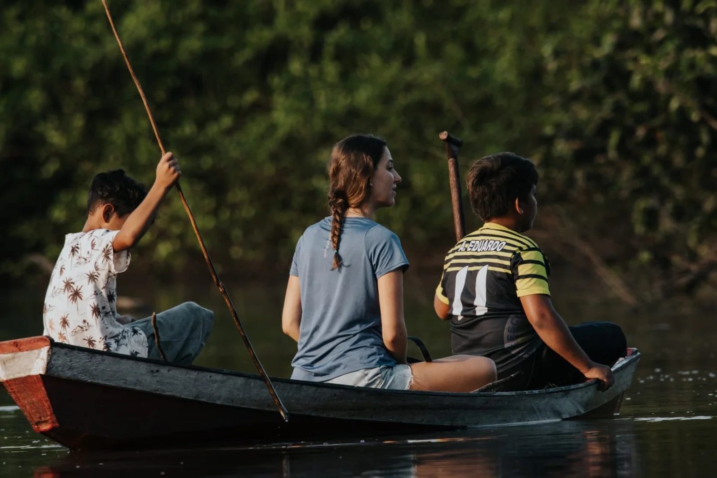 Three paddlers, one canoe, a thousand shades of green. A woman and two local boys paddle a wooden canoe along a calm jungle river during an Amazon trek and survival tour, dense green forest lining the banks