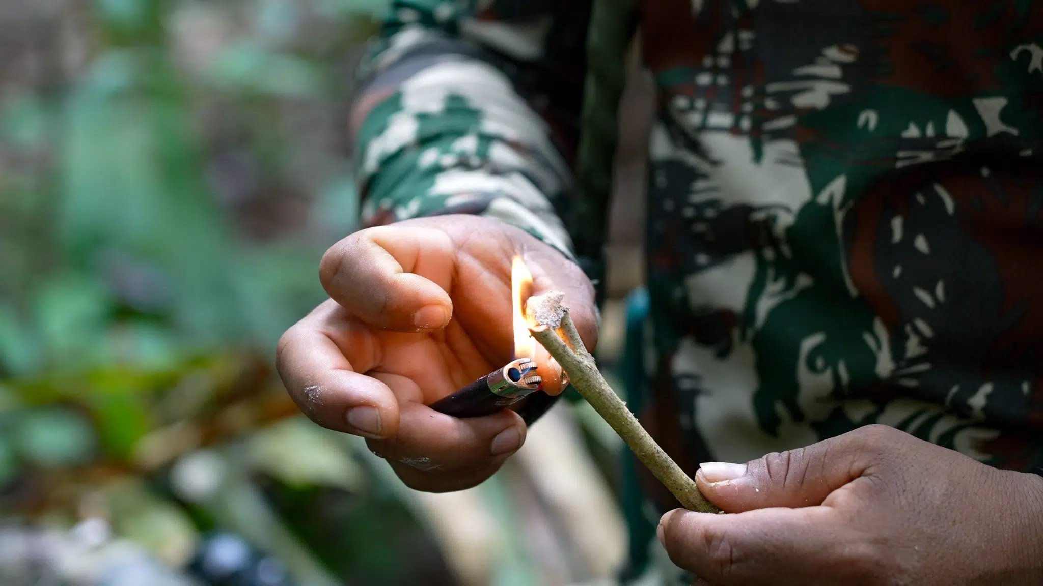 Flame conjured from a single stick — survival knowledge passed hand to hand. A guide in camouflage clothing lights a green stick using a lighter during an Amazon trek and survival tour, demonstrating a bushcraft fire-starting technique in the rainforest