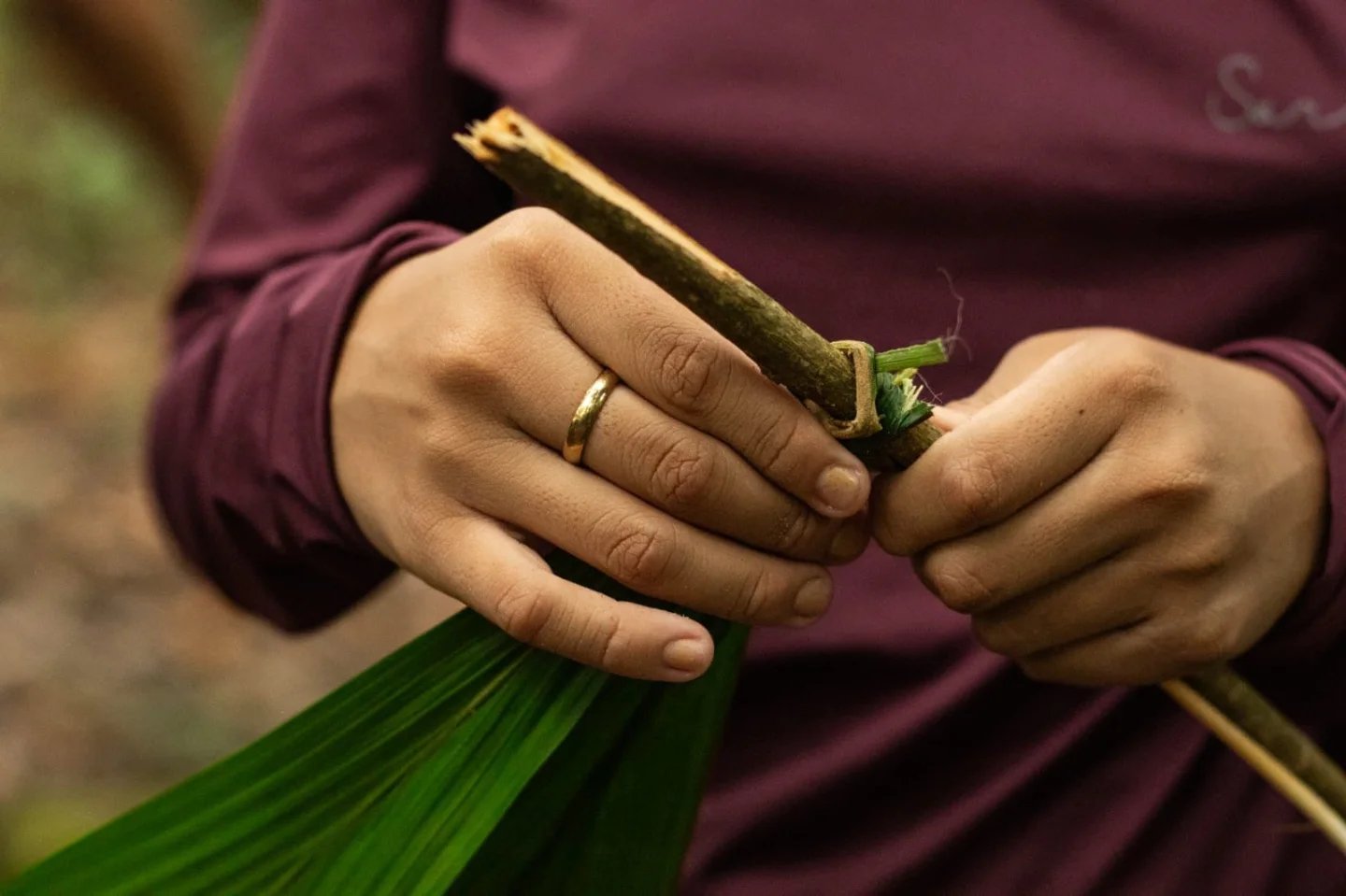 Lashing leaves to wood — survival knowledge passed hand to hand. Close-up of hands binding a wooden stick to green palm fronds during an Amazon trek and survival tour jungle craft demonstration