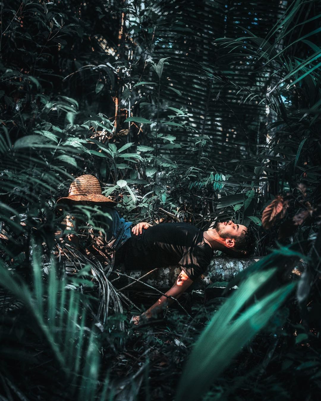 Stillness earned — the forest floor reclaims you. A man lies back on a mossy log deep in dense rainforest undergrowth, his straw hat set aside, resting during an Amazon trek and survival tour