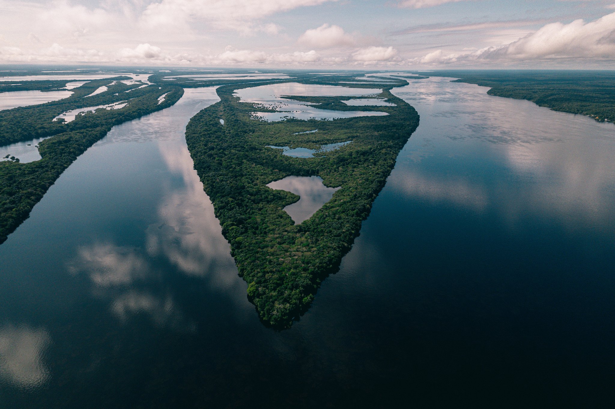 Hundreds of islands, one unbroken canopy — the Amazon at its most elemental. Aerial view of a densely forested island at Anavilhanas Jungle Lodge Brazil, surrounded by the dark mirror-calm waters of a vast river archipelago under a cloudy sky