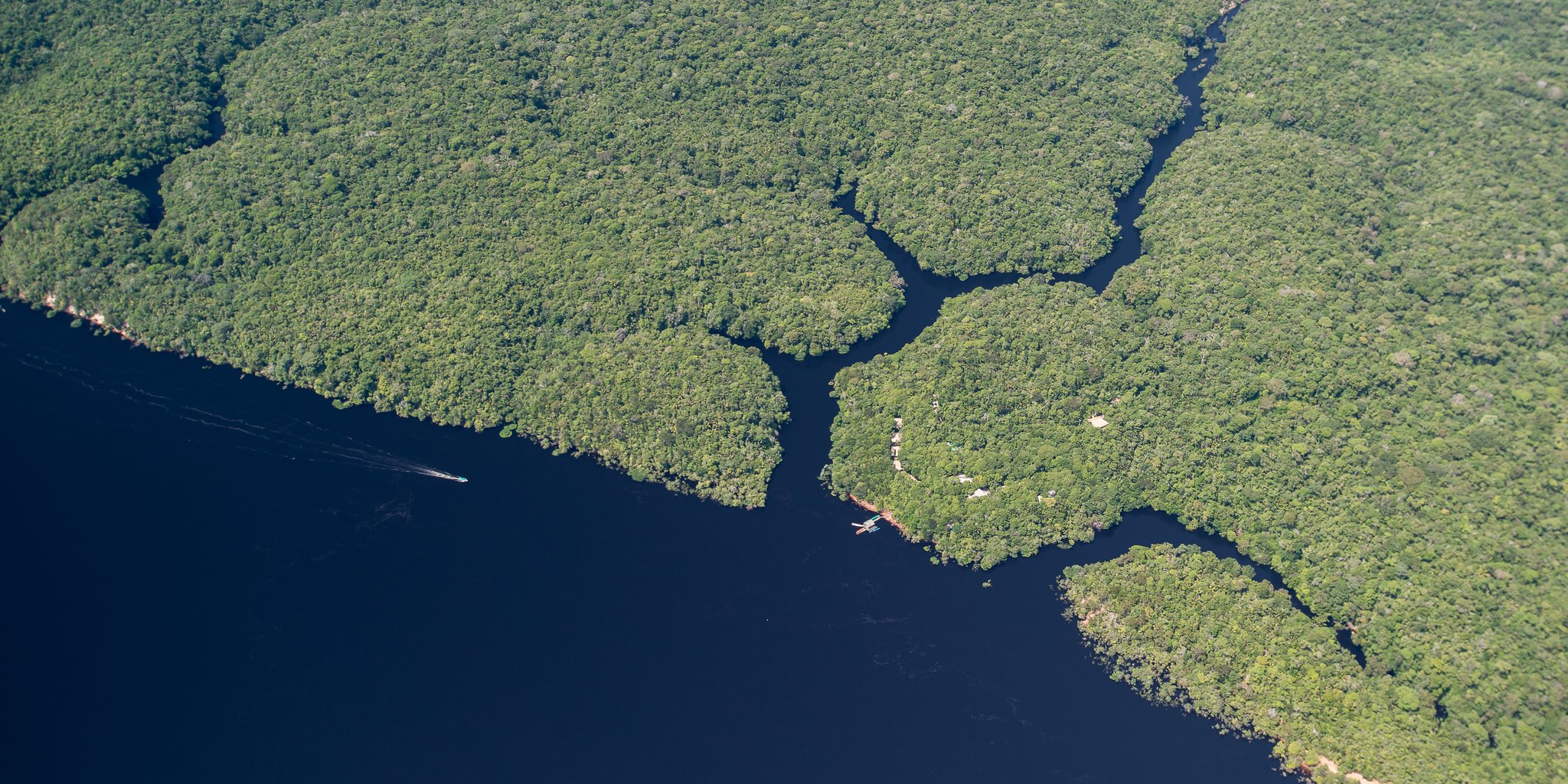 Tiny against the vastness — the lodge, the channels, the endless green. Photo: MARCELO ISOLA Aerial view of Anavilhanas Jungle Lodge Brazil nestled at the edge of dark Rio Negro waters, surrounded by an unbroken carpet of dense Amazon rainforest canopy with narrow channels threading between forested islands, a small boat leaving a wake on the river below