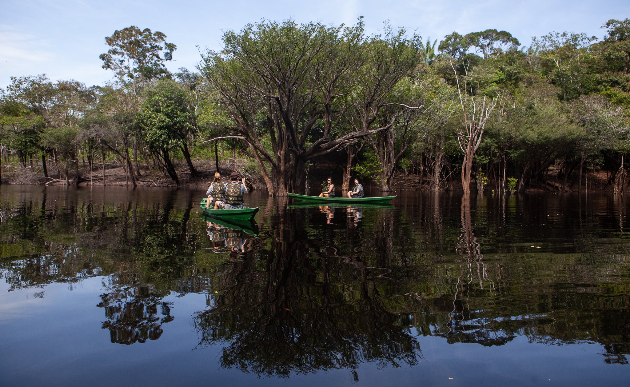 Green canoe, black water, trees rising straight from the flood. Three travellers in a green canoe gliding through a flooded forest on dark mirror-still water at Anavilhanas Jungle Lodge Brazil, surrounded by partially submerged trees and their perfect reflections
