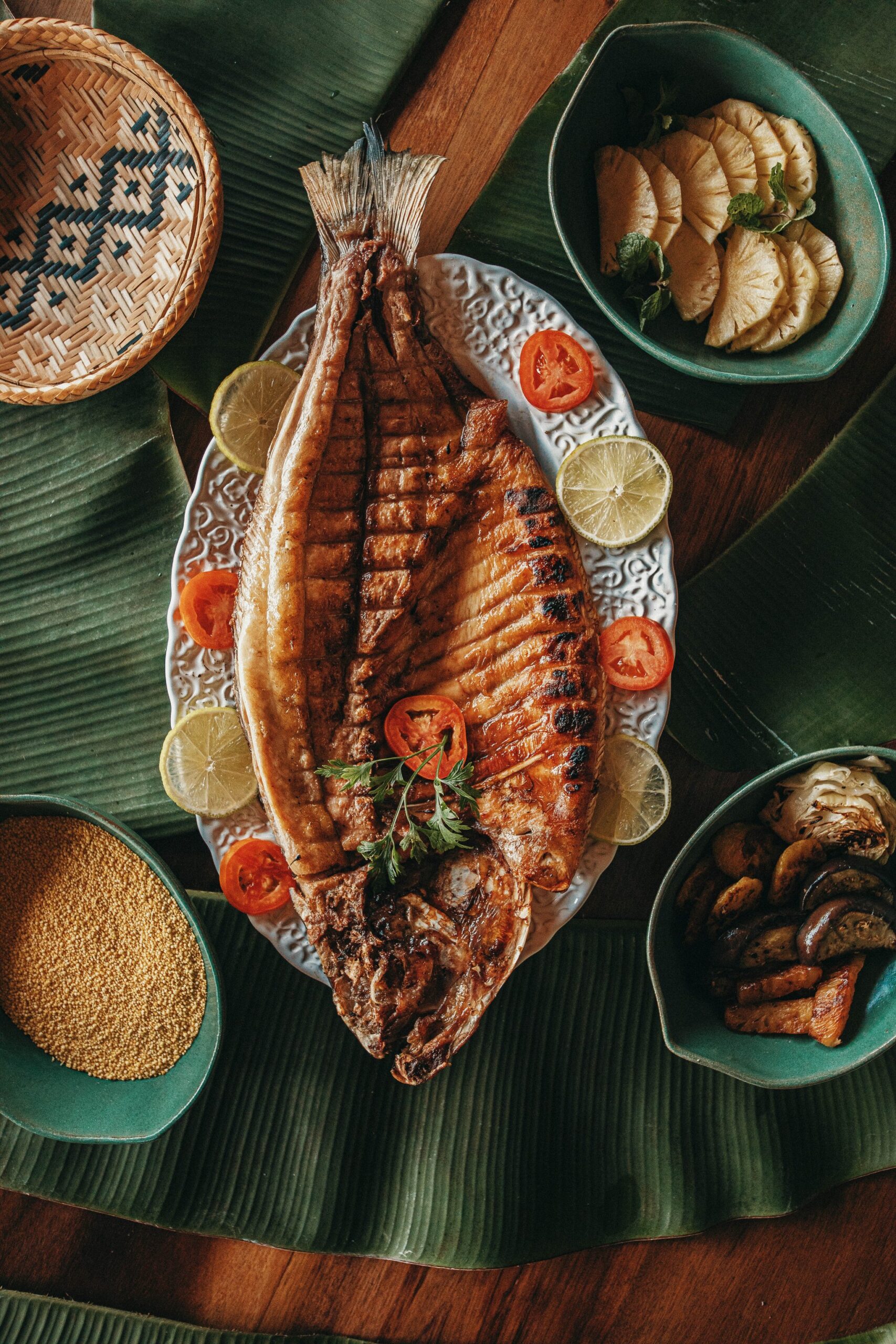 Char marks, citrus, and the Amazon on your plate. A whole grilled fish served on a decorative white platter with lime and tomato slices at Anavilhanas Jungle Lodge Brazil, surrounded by side bowls of pineapple, roasted mushrooms and golden seeds on banana leaves