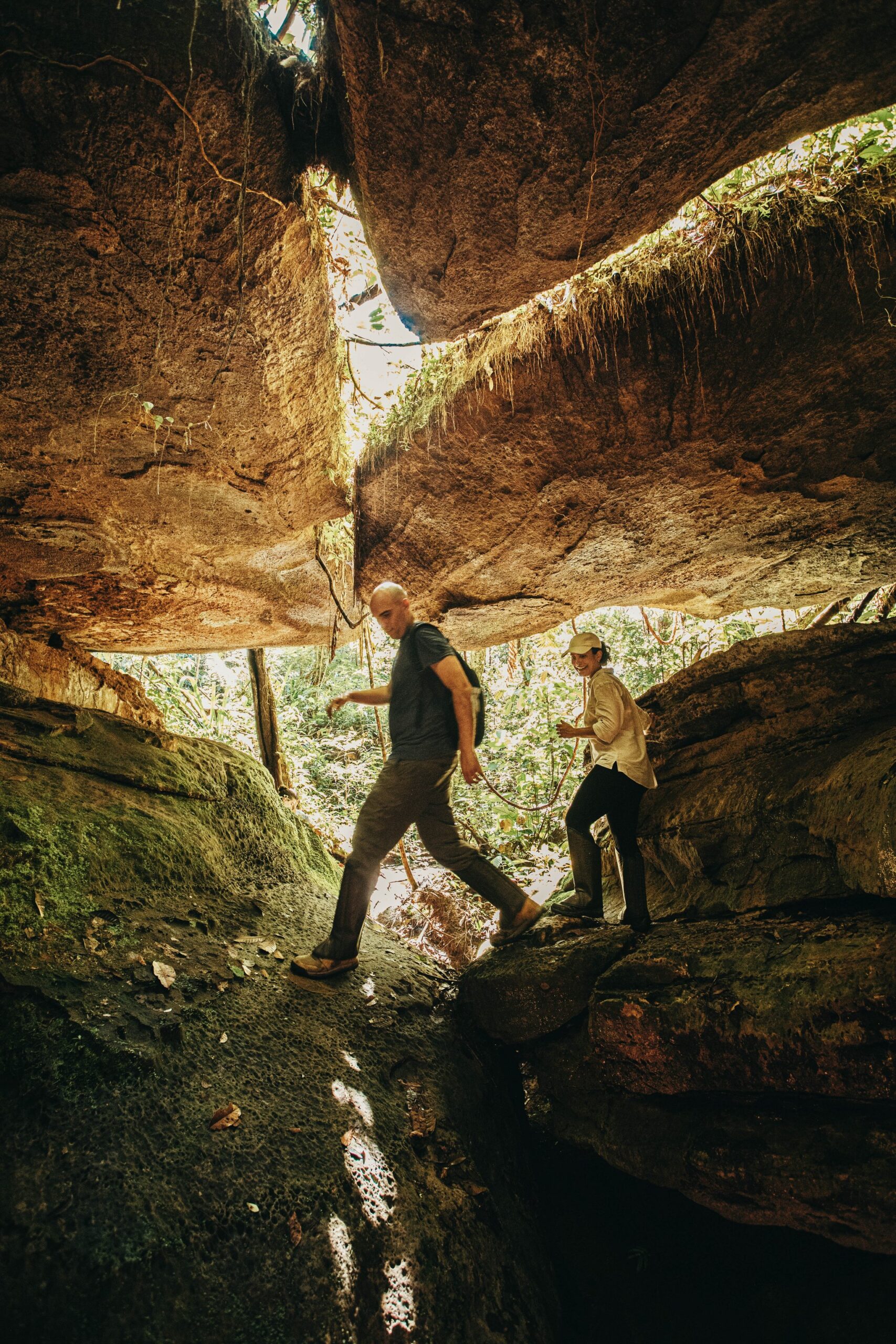 Ducking through 700-million-year-old rock, the jungle blazing bright at every gap. Two hikers duck beneath a massive sandstone overhang at the Madadá Caves during an Anavilhanas Jungle Lodge Brazil excursion, sunlit jungle visible through the rock opening behind them
