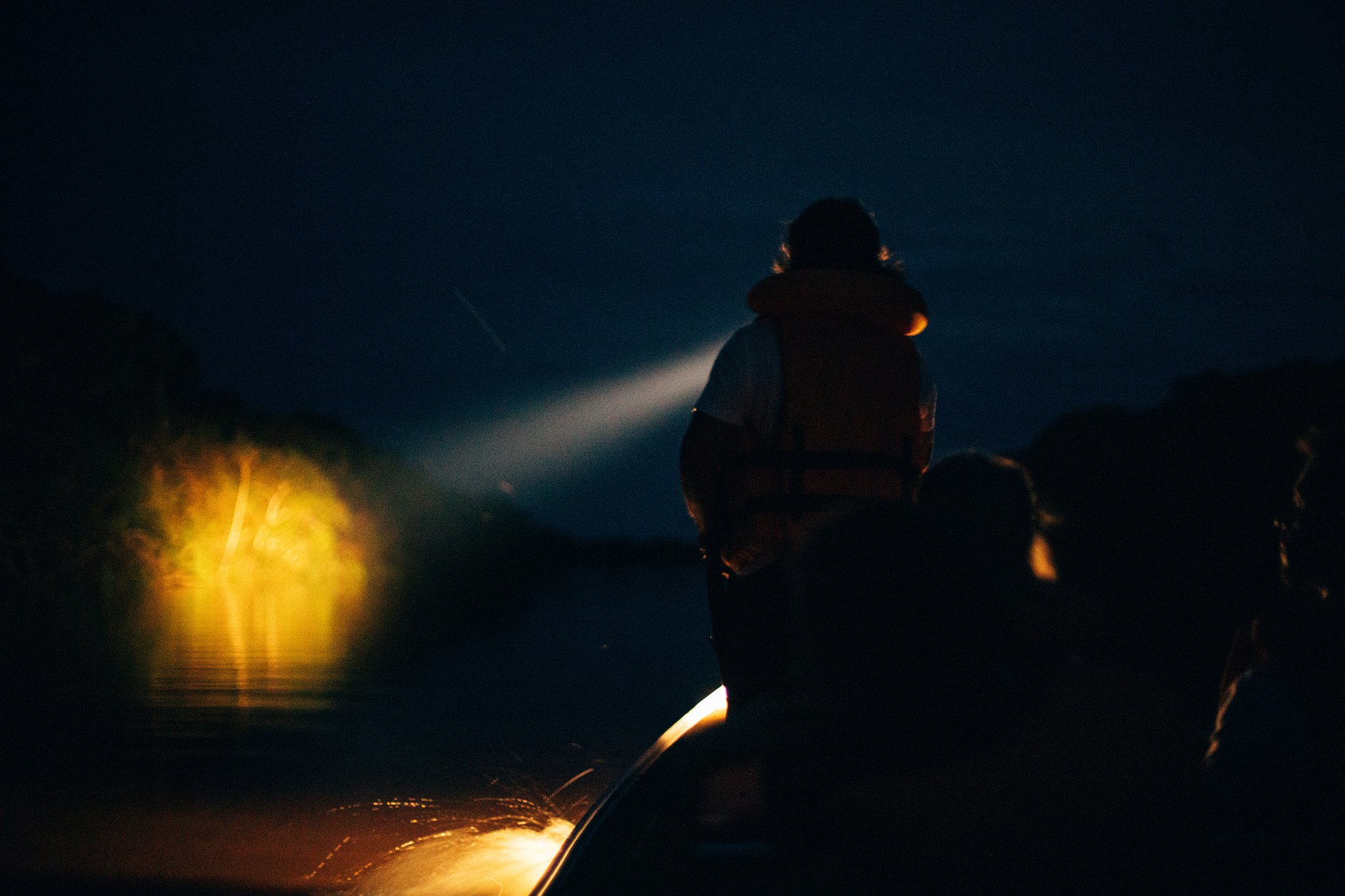 Spotlight cutting through the dark — the Amazon reveals itself after nightfall. A silhouetted guide in a life vest sweeps a spotlight across dark river water during a night tour at Anavilhanas Jungle Lodge Brazil, with a warm yellow lamp glowing at the bow