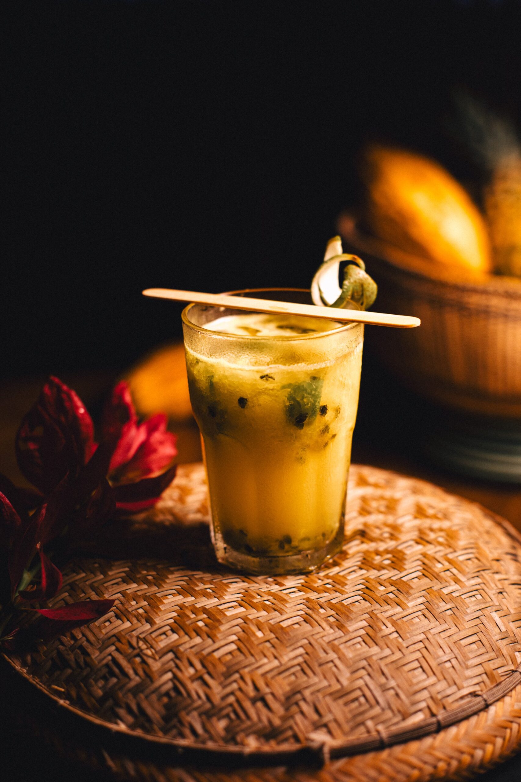 Tangy, golden, and impossibly refreshing — passion fruit straight from the Amazon. A golden passion fruit cocktail with visible seeds served in a glass on a woven rattan tray at Anavilhanas Jungle Lodge Brazil, garnished with a wooden stirrer and a curled tropical leaf, with a basket of yellow fruit softly lit in the background