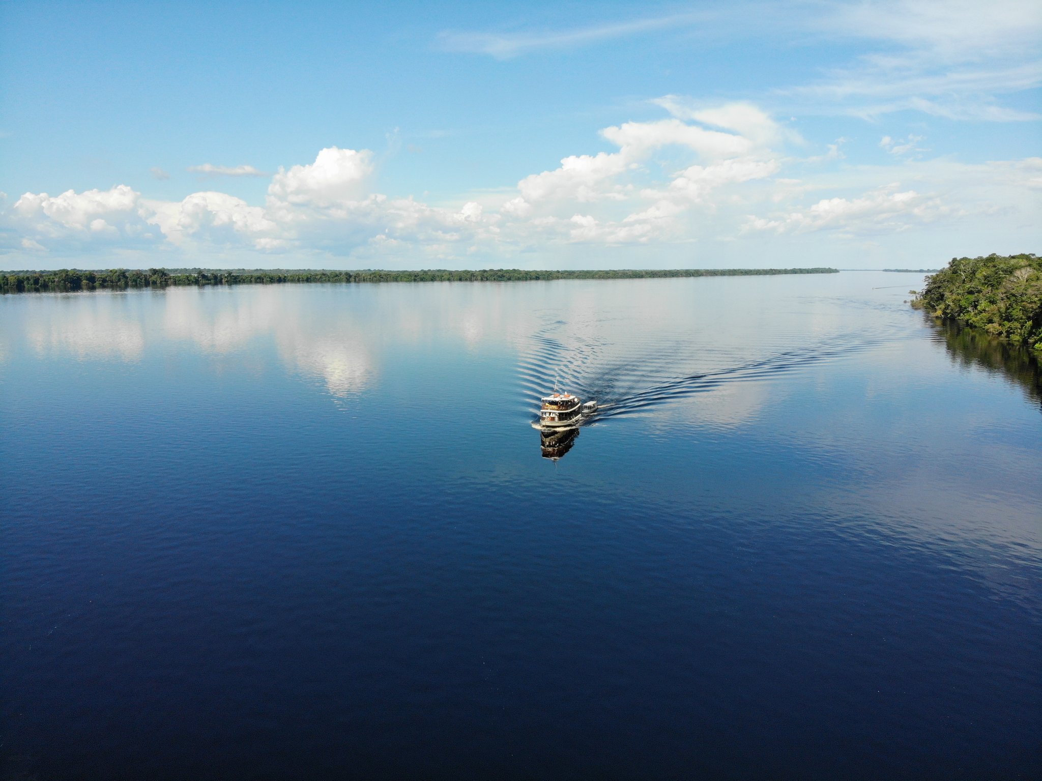 Aerial view of a small expedition vessel crossing the glassy dark waters of the Rio Negro, flanked by dense Amazon rainforest on the horizon, on a journey to Anavilhanas Jungle Lodge Brazil