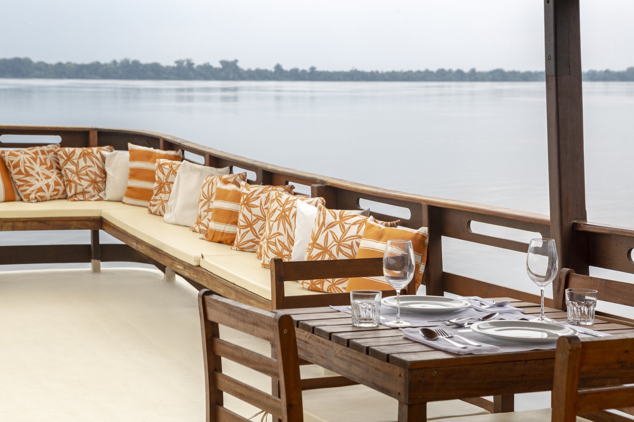 Lunch served on the water, forest at the edges of everything. A set dining table with wine glasses and cutlery on the open deck of a boat at Anavilhanas Jungle Lodge Brazil, with a cushioned lounge bench and a wide river stretching to the forested horizon