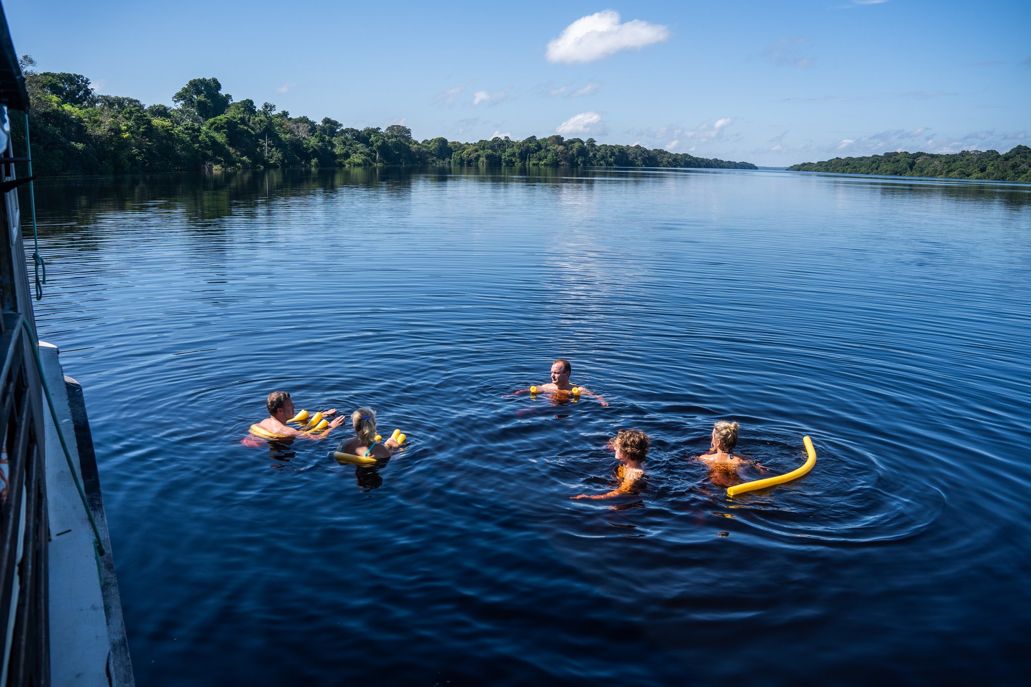 Cool, dark water and nothing but forest for miles. Photo: Alex Fisberg Five guests swimming in the dark waters of the Rio Negro beside a traditional boat at Anavilhanas Jungle Lodge Brazil, with yellow foam noodles and dense rainforest lining both riverbanks under a blue sky